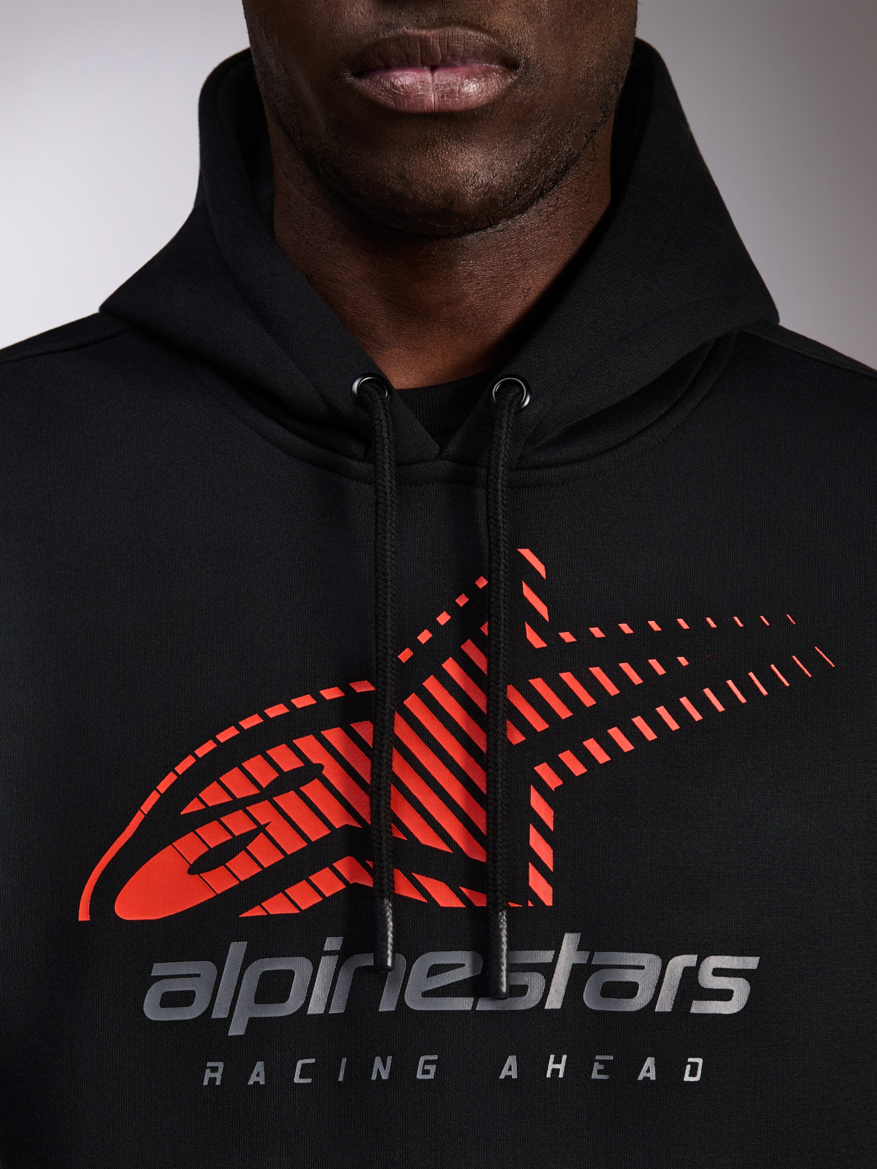Alpinestars Pulse Hoodie, sudadera negra con capucha, primer plano del pecho con logotipo Astars rojo con degradado de líneas de velocidad y marca plateada, cordones negros con puntas sumergidas, estilo streetwear casual de motor