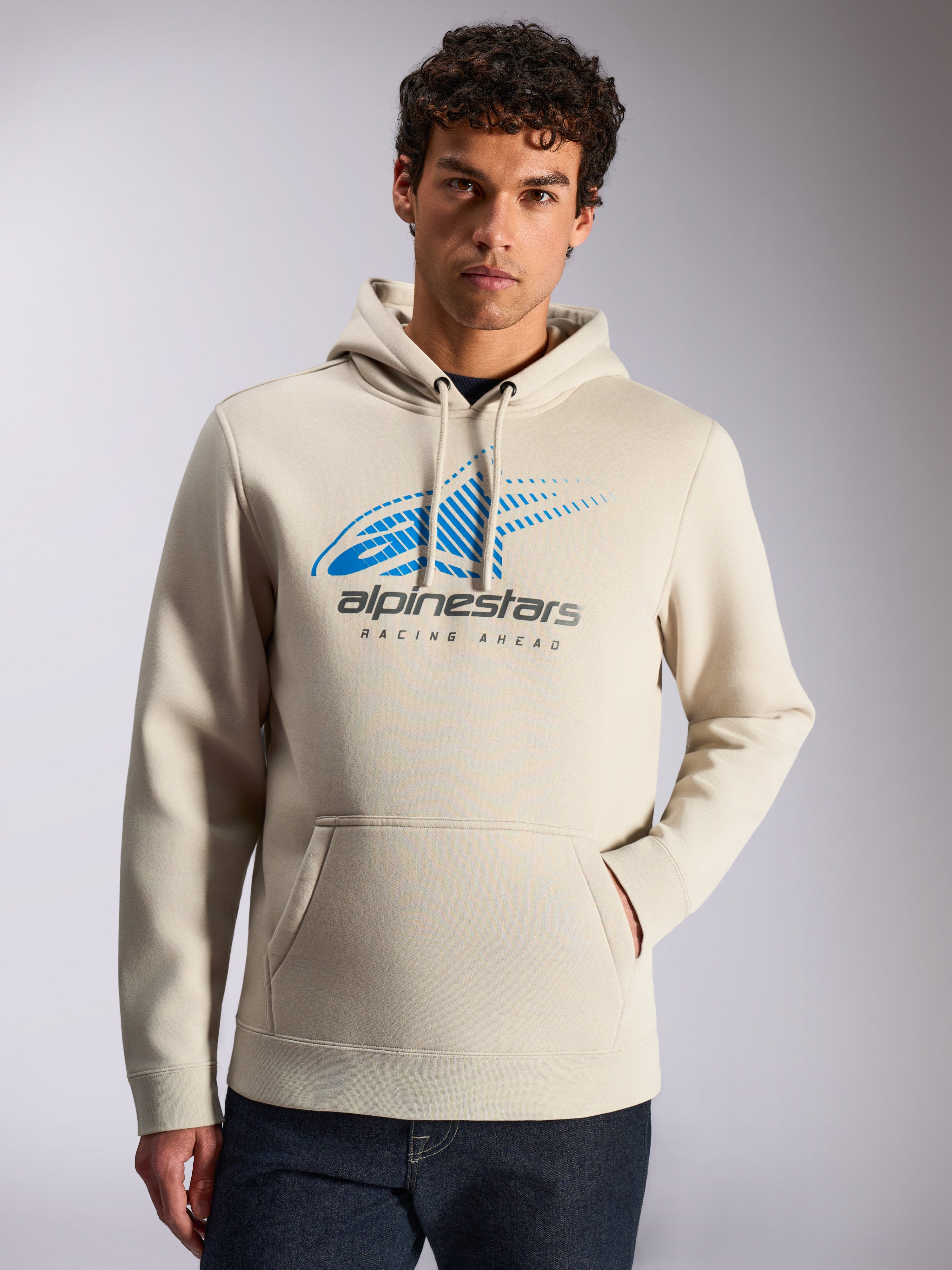 Alpinestars Pulse Hoodie, ropa urbana casual, color arena, logo frontal Astars Combo en azul y gris con desvanecimiento de líneas de velocidad y texto 'Racing Ahead', capucha con cordón, bolsillo de canguro, usada por un modelo con jeans oscuros