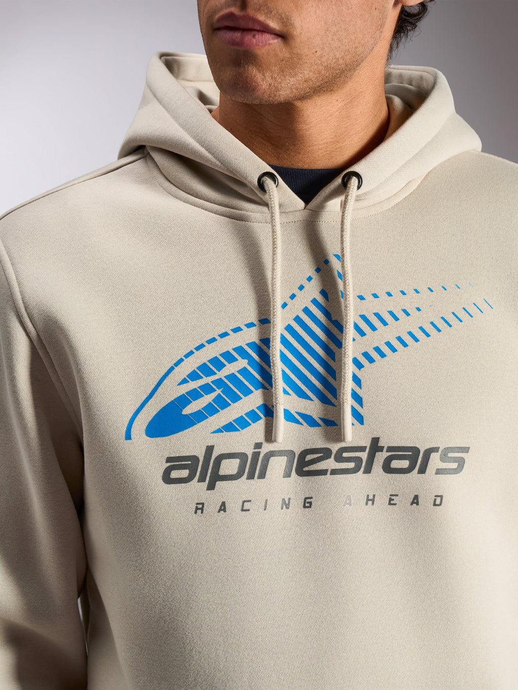 Alpinestars Pulse Hoodie, sudadera informal tipo pullover en color Off White, primer plano del pecho con logo Astars Combo azul y gris con líneas de velocidad y texto 'Racing Ahead', capucha con cordón y puntas sumergidas.