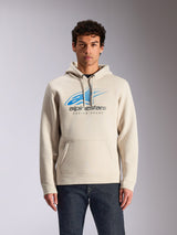 Sudadera con capucha Alpinestars Pulse, sudadera informal tipo pullover en color blanco roto, logotipo frontal Astars Combo en azul y gris con gráfico de líneas de velocidad degradadas, bolsillo de canguro frontal, vestida por un modelo con vaqueros oscuros, moda urbana inspirada en el motociclismo