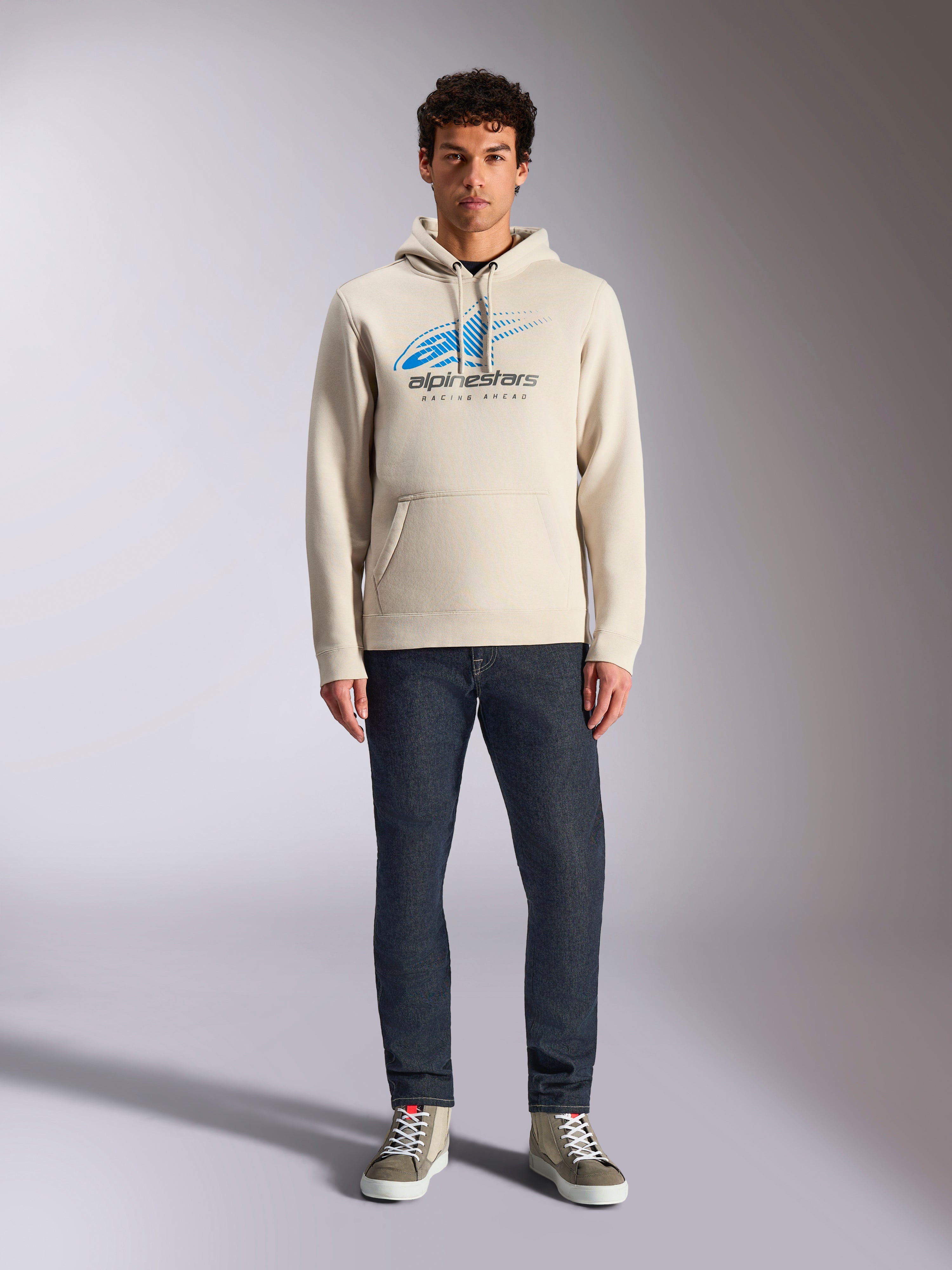 Alpinestars Pulse Hoodie, Off White, sudadera de manga larga con capucha y cordón, logotipo Astars Combo en azul y negro con líneas de velocidad desvanecidas, combinada con vaqueros oscuros y zapatillas, estilo casual motorsport streetwear