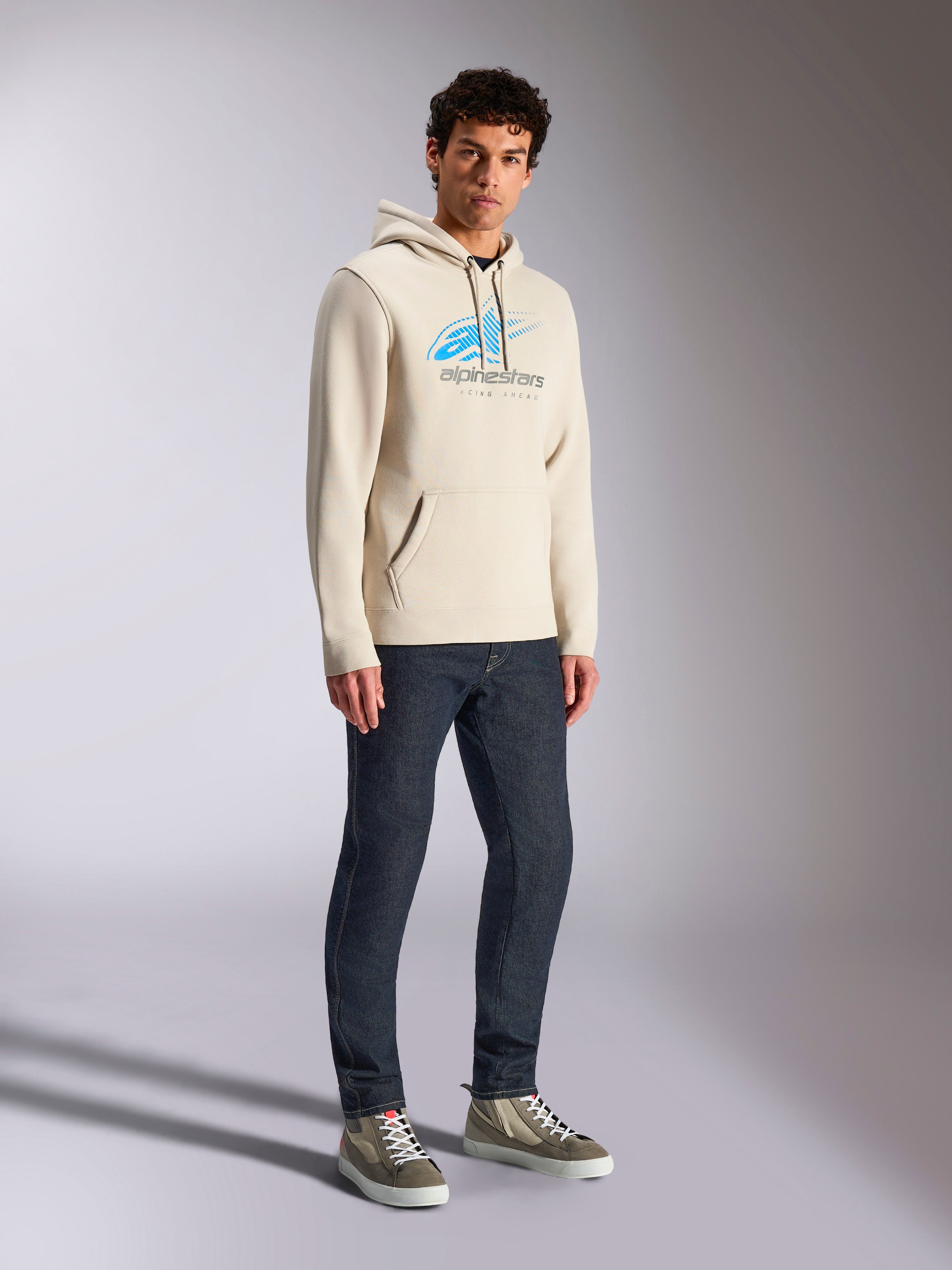 Sudadera con capucha Alpinestars Pulse, ropa urbana casual en color Off White, sudadera de felpa con logo Astars frontal en azul y gris con líneas de velocidad desvanecidas, bolsillo de canguro, lucida por un modelo con vaqueros oscuros y zapatillas de caña alta.