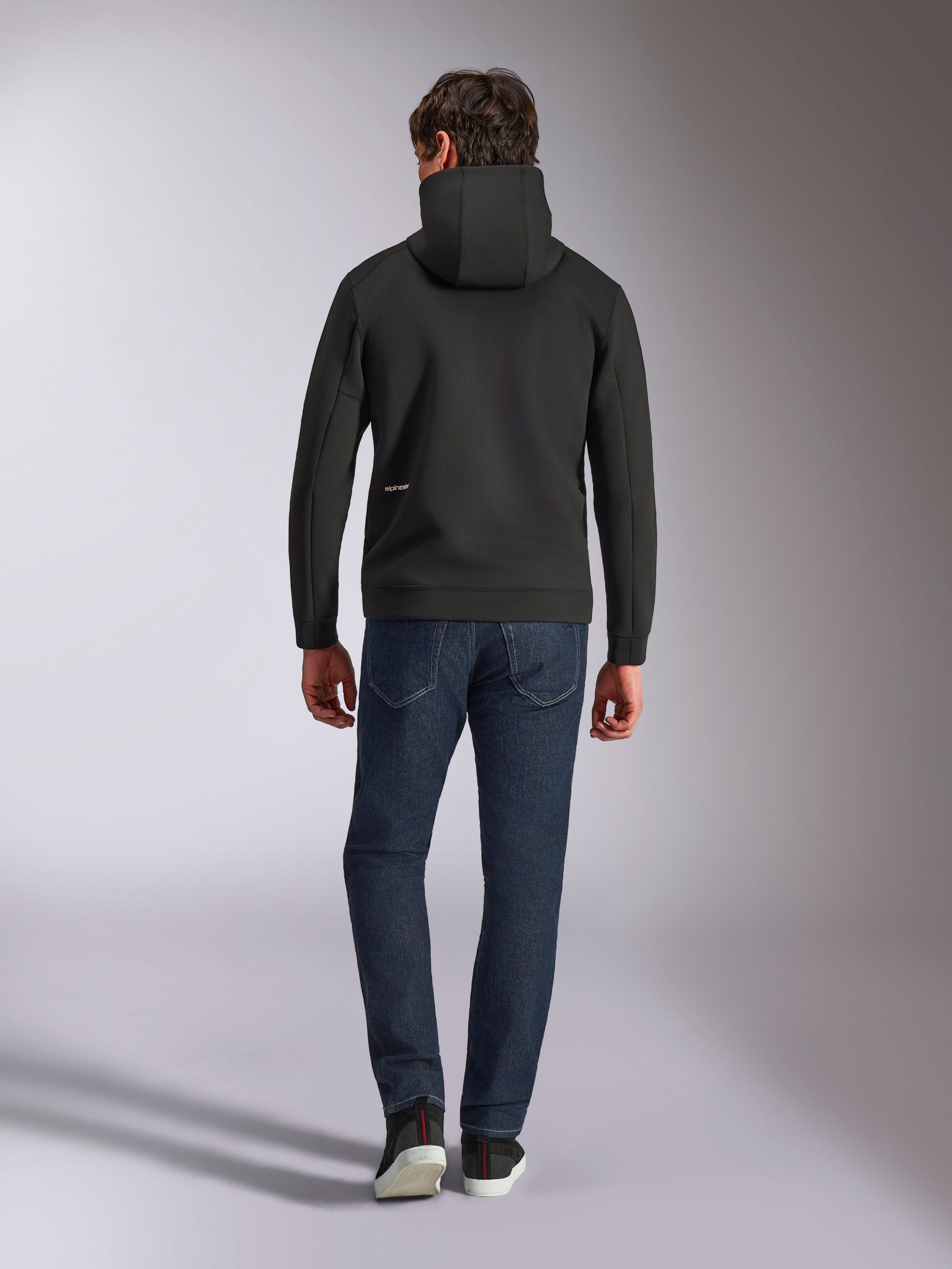 Alpinestars Cypher Hoodie, sudadera de forro polar negro de doble cara, vista posterior con un pequeño logotipo blanco de Alpinestars en la cadera izquierda, capucha de cuello alto, combinada con vaqueros azul oscuro, estilo casual inspirado en el automovilismo
