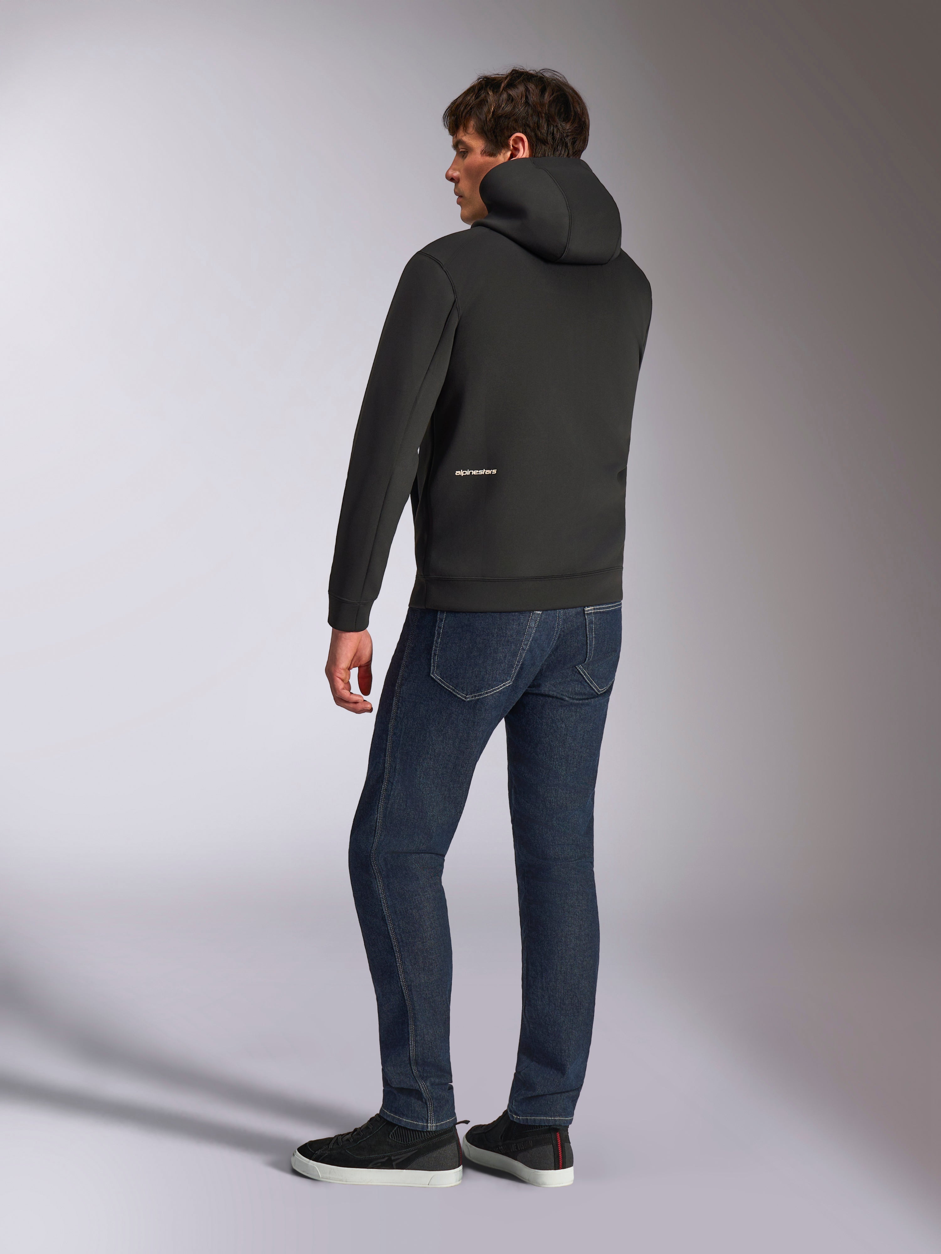 Sudadera con capucha Cypher de Alpinestars, sudadera casual negra con cremallera, vista trasera que muestra la capucha de cuello alto y el logotipo de Alpinestars en blanco en la parte inferior de la espalda, combinada con vaqueros azul oscuro y zapatillas negras, diseño moderno inspirado en el automovilismo