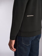 Sudadera con capucha Alpinestars Cypher, vista de primer plano de la parte inferior de la espalda y la manga, forro polar negro de doble cara con el logotipo de Alpinestars en blanco, combinada con vaqueros azules, ropa urbana informal de estilo motorsport