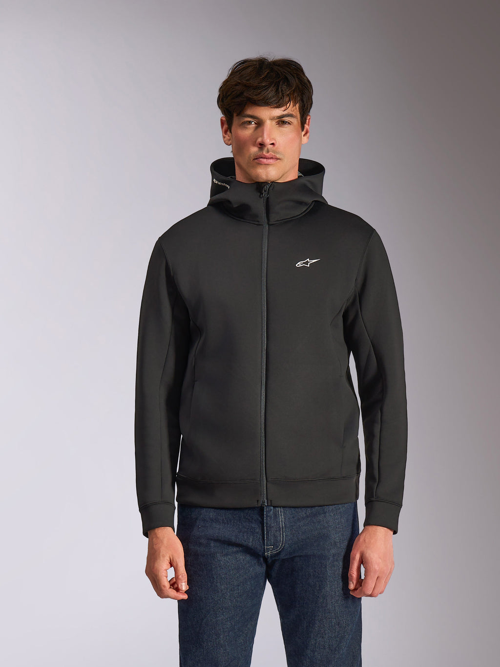 Sudadera con capucha Alpinestars Cypher, sudadera de forro polar negra con cremallera frontal, con capucha de cuello alto con marca blanca y un pequeño logo Astars blanco en el pecho, usada por un modelo con vaqueros azules, ropa urbana casual inspirada en el motorsport.