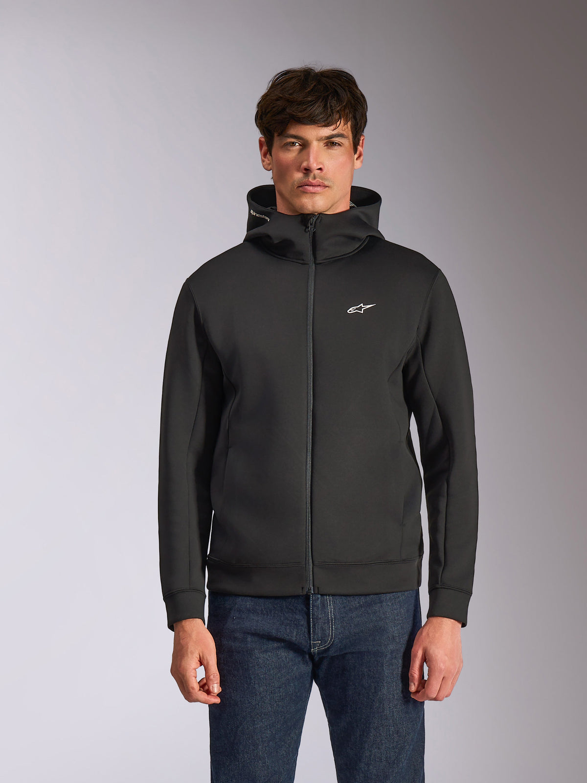 Sudadera con capucha Alpinestars Cypher, sudadera de forro polar negra con cremallera frontal, con capucha de cuello alto con marca blanca y un pequeño logo Astars blanco en el pecho, usada por un modelo con vaqueros azules, ropa urbana casual inspirada en el motorsport.
