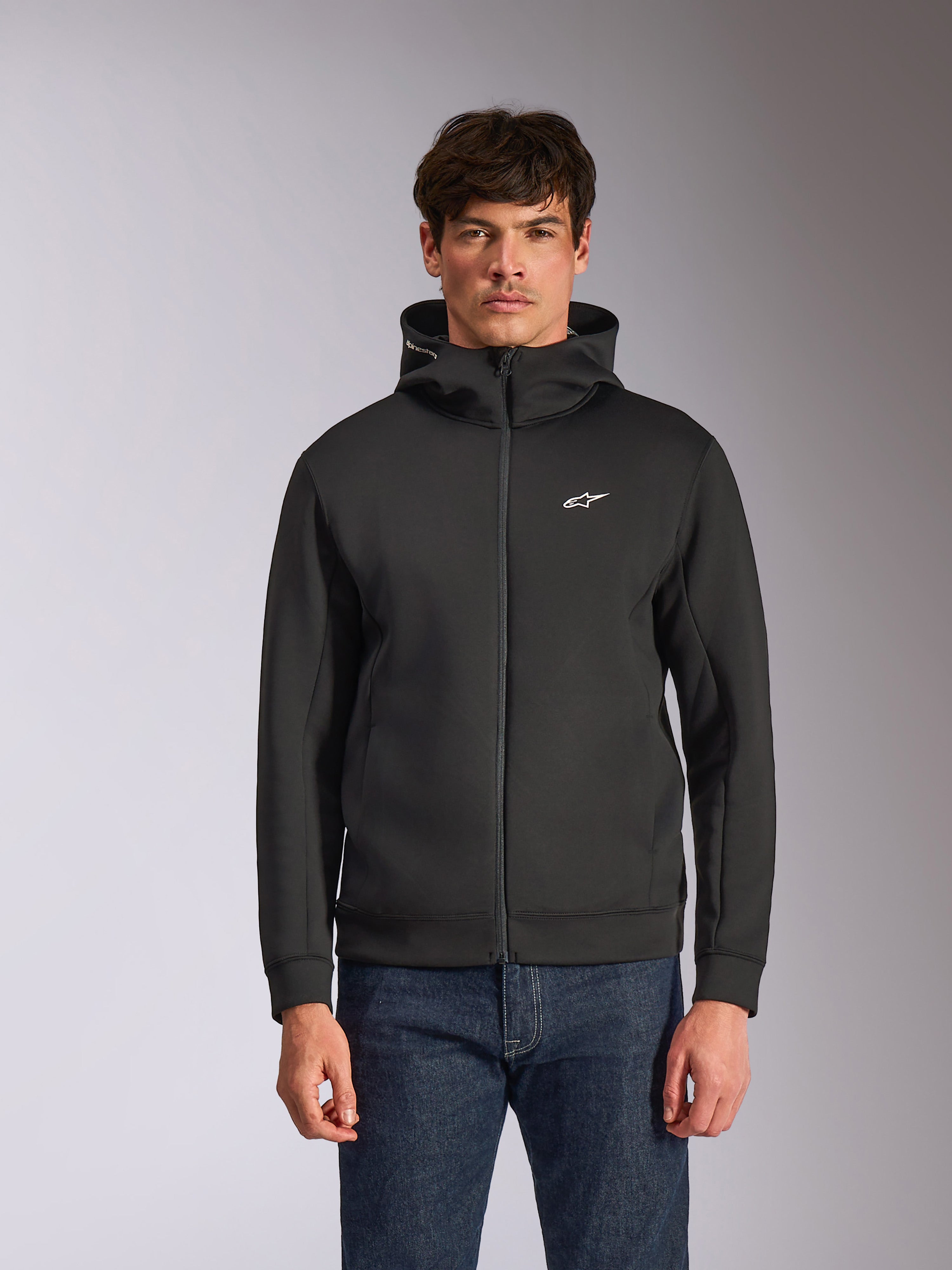 Sudadera con capucha Alpinestars Cypher, sudadera de forro polar negra con cremallera frontal, con capucha de cuello alto con marca blanca y un pequeño logo Astars blanco en el pecho, usada por un modelo con vaqueros azules, ropa urbana casual inspirada en el motorsport.