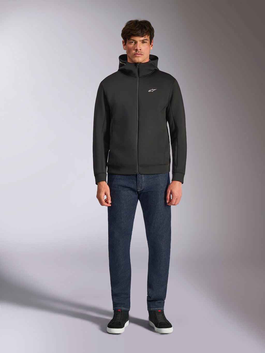 Modelo con la sudadera Alpinestars Cypher Hoodie, sudadera de forro polar negra con cremallera frontal, capucha de cuello alto con pequeño logo Astars blanco en el pecho, combinada con vaqueros de denim oscuro y zapatillas negras, ropa urbana casual inspirada en el automovilismo