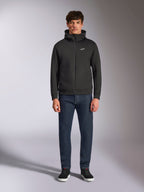 Modelo con la sudadera Alpinestars Cypher Hoodie, sudadera de forro polar negra con cremallera frontal, capucha de cuello alto con pequeño logo Astars blanco en el pecho, combinada con vaqueros de denim oscuro y zapatillas negras, ropa urbana casual inspirada en el automovilismo