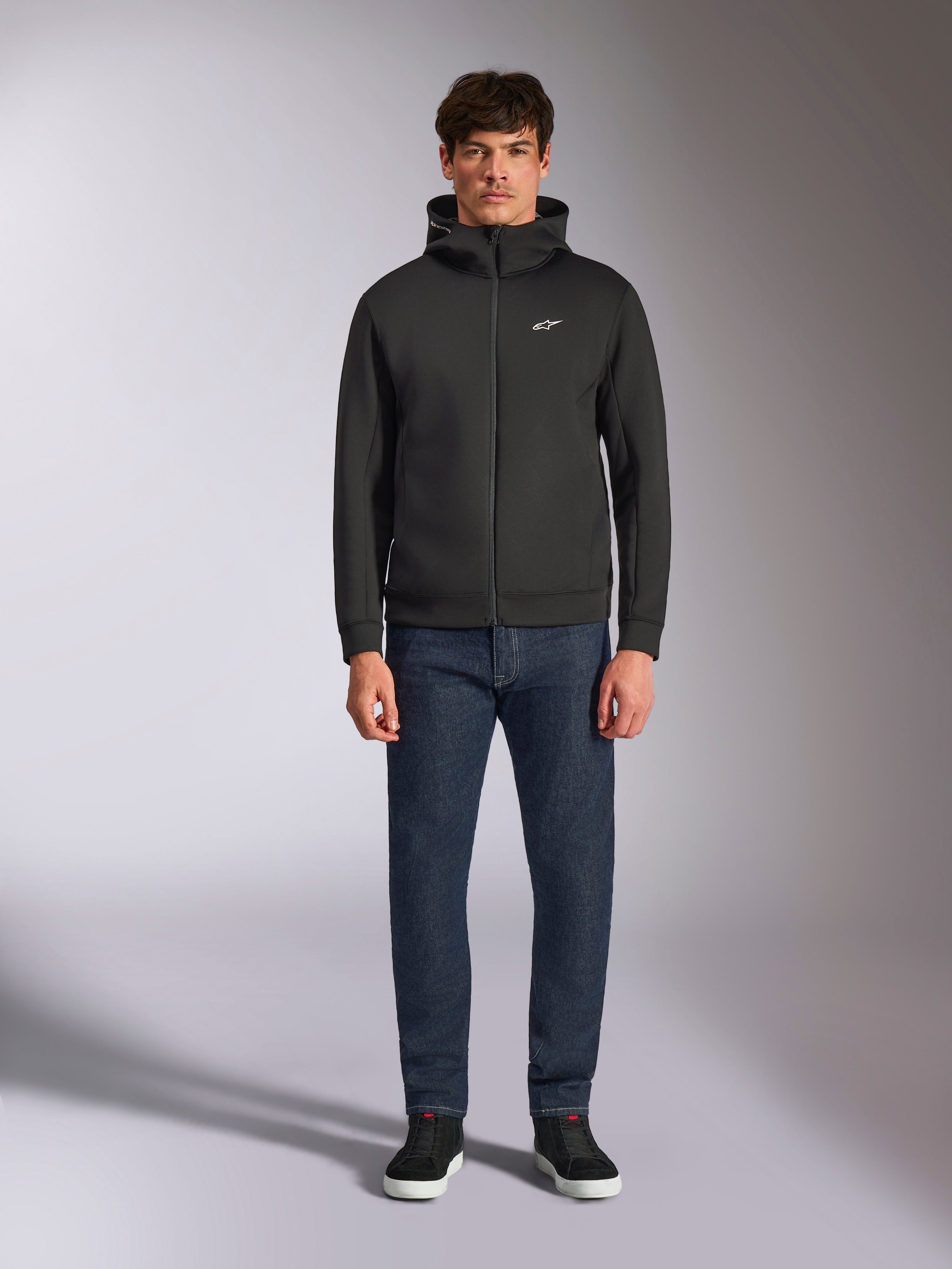 Modelo con la sudadera Alpinestars Cypher Hoodie, sudadera de forro polar negra con cremallera frontal, capucha de cuello alto con pequeño logo Astars blanco en el pecho, combinada con vaqueros de denim oscuro y zapatillas negras, ropa urbana casual inspirada en el automovilismo