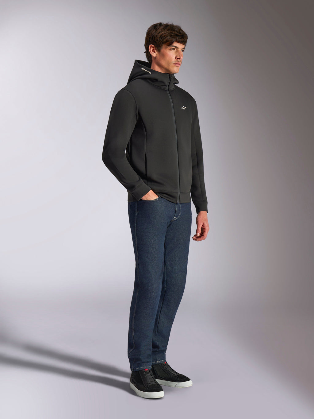 Sudadera con capucha Alpinestars Cypher, ropa urbana casual, sudadera negra con cremallera, modelo masculino con pantalones vaqueros azul oscuro y zapatillas negras, capucha de cuello alto con logotipo de Alpinestars, pequeño logo Astars blanco en el pecho, look estilizado inspirado en el motociclismo