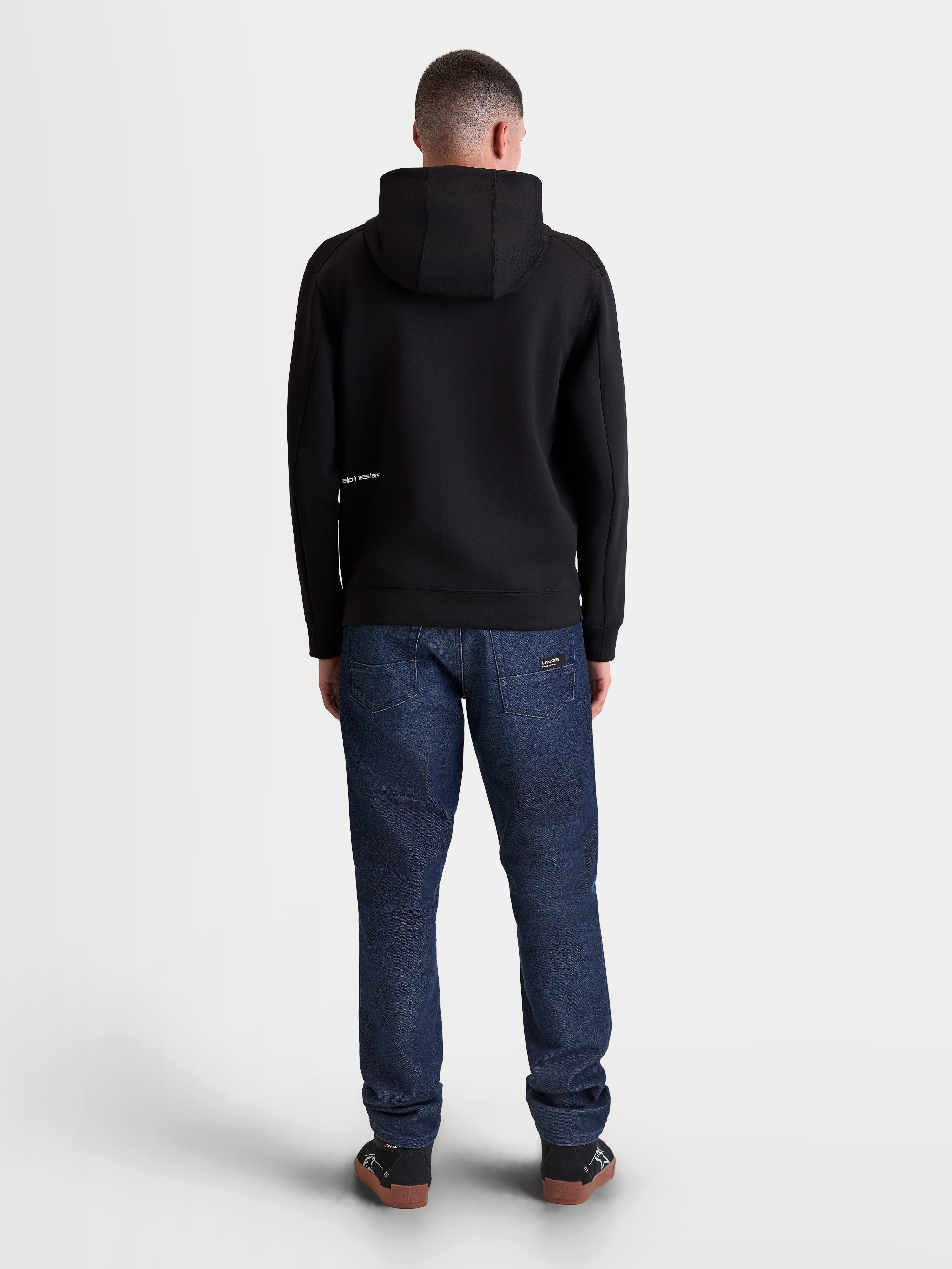 Alpinestars sudadera con capucha Cypher, sudadera de forro polar premium en color negro con cremallera frontal, vista posterior en modelo mostrando la capucha de cuello alto y detalle de logotipo minimalista, tejido técnico de mezcla triple, ropa deportiva casual y estilo de vida motorsports.