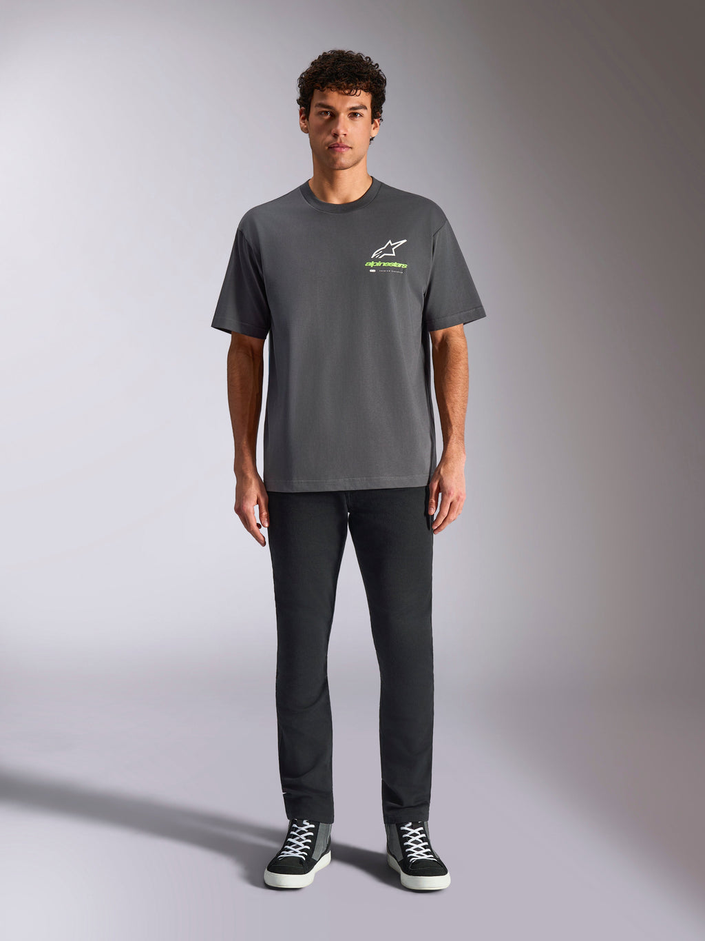 Camiseta Alpinestars Overhang Oversized CSF, camiseta de manga corta, gris carbón, con logotipo frontal de Alpinestars en blanco y verde lima, vestida por un modelo con pantalones negros y zapatillas, estilo casual streetwear relajado