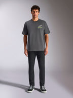 Camiseta Alpinestars Overhang Oversized CSF, camiseta de manga corta, gris carbón, con logotipo frontal de Alpinestars en blanco y verde lima, vestida por un modelo con pantalones negros y zapatillas, estilo casual streetwear relajado