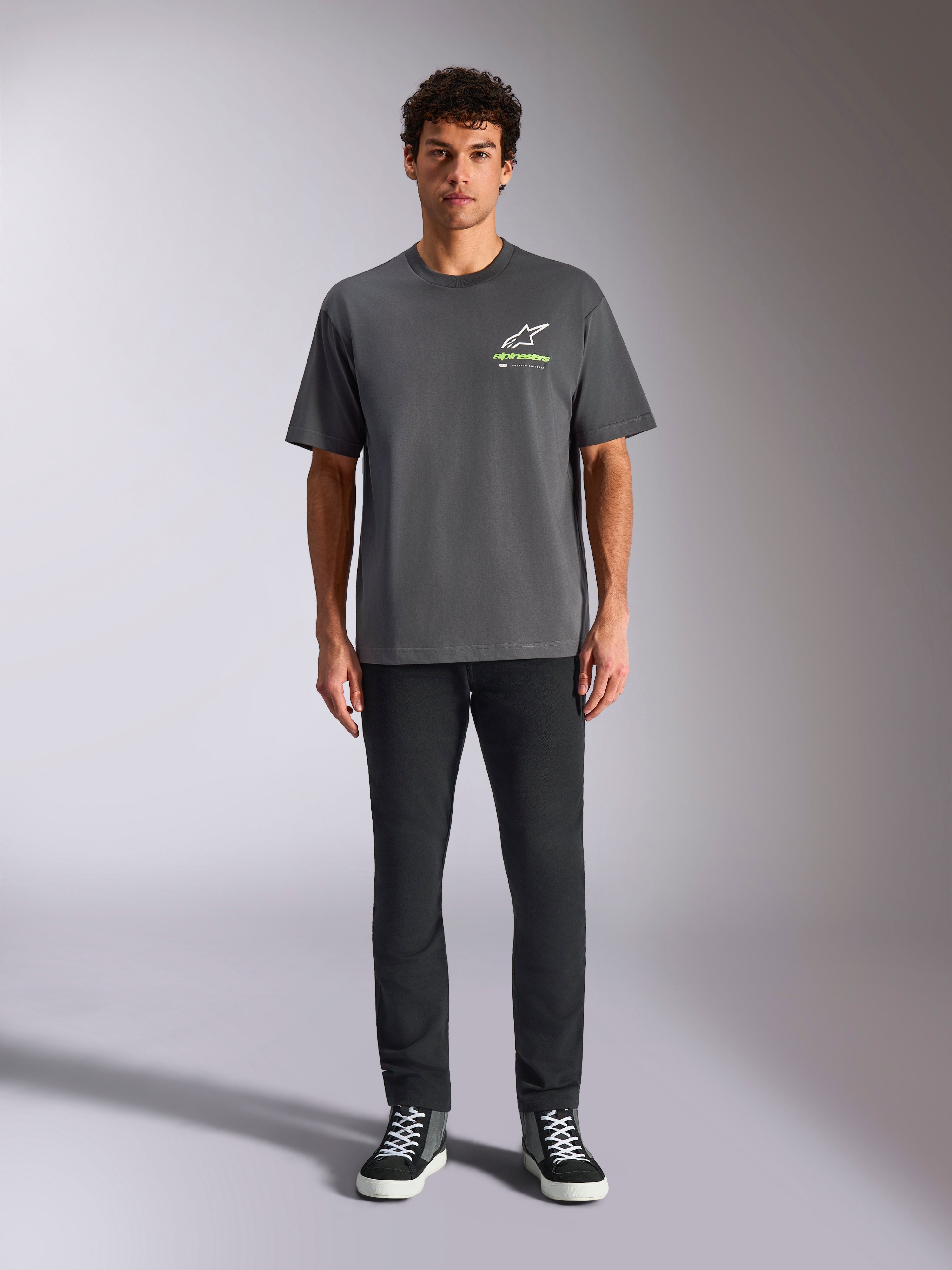 Camiseta Alpinestars Overhang Oversized CSF, camiseta de manga corta, gris carbón, con logotipo frontal de Alpinestars en blanco y verde lima, vestida por un modelo con pantalones negros y zapatillas, estilo casual streetwear relajado