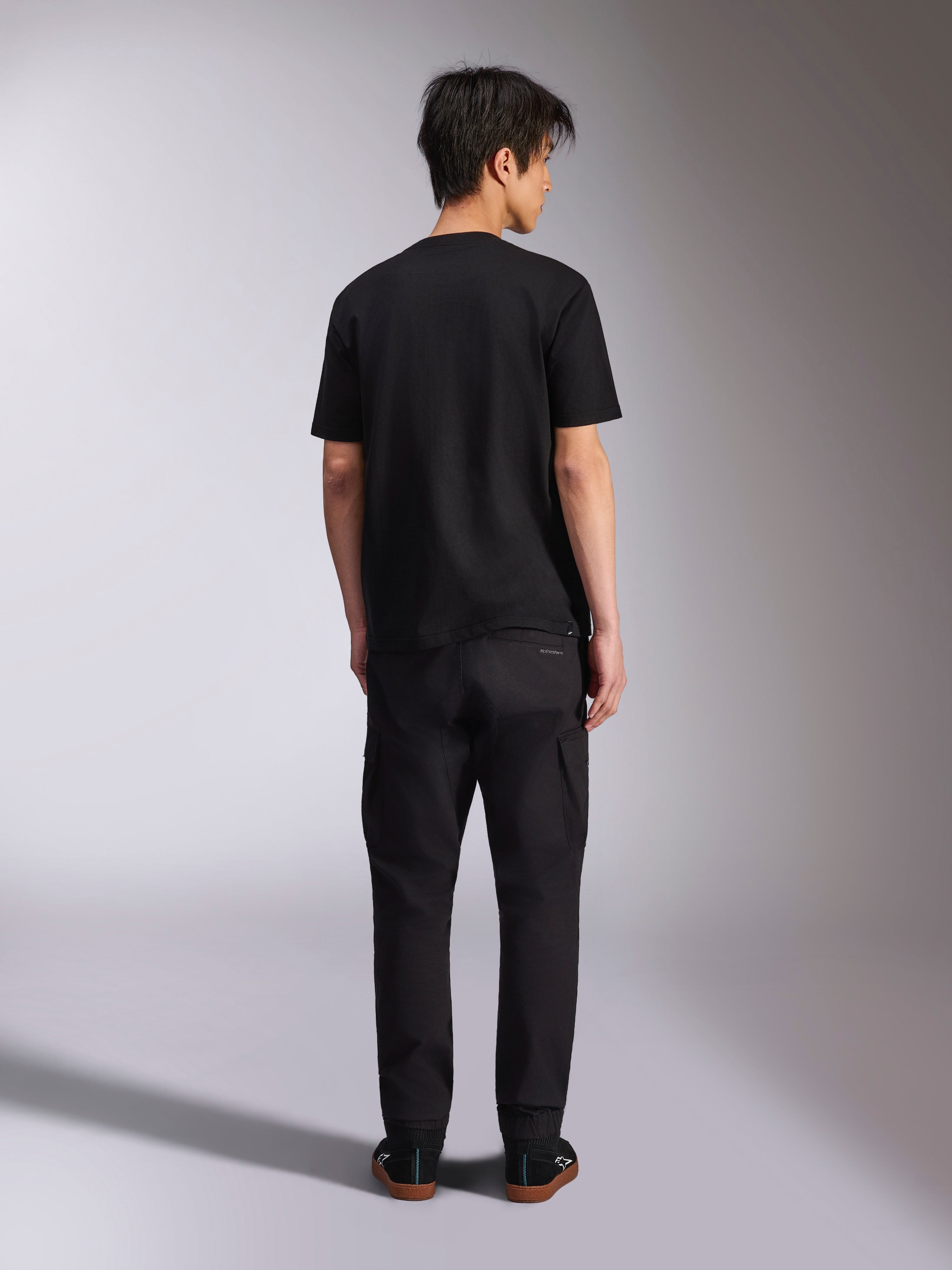 Alpinestars Noka CSF Tee, camiseta de manga corta, negra, vista trasera del modelo vistiendo la camiseta con pantalones negros y zapatillas, estilo streetwear casual de motorsport