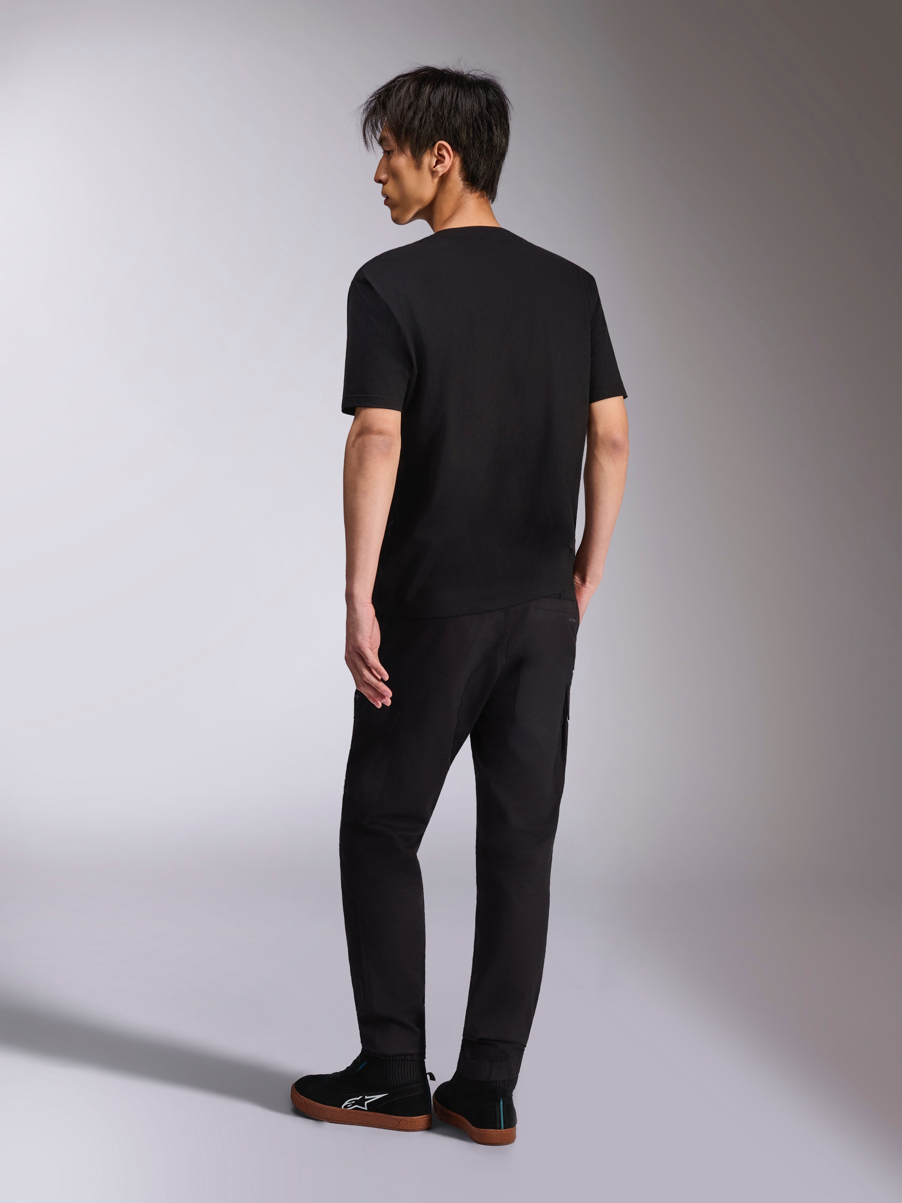 Alpinestars Noka CSF Tee, camiseta de manga corta, negra, vista posterior en modelo, combinada con pantalones negros y zapatillas casuales, estilo urbano minimalista de automovilismo