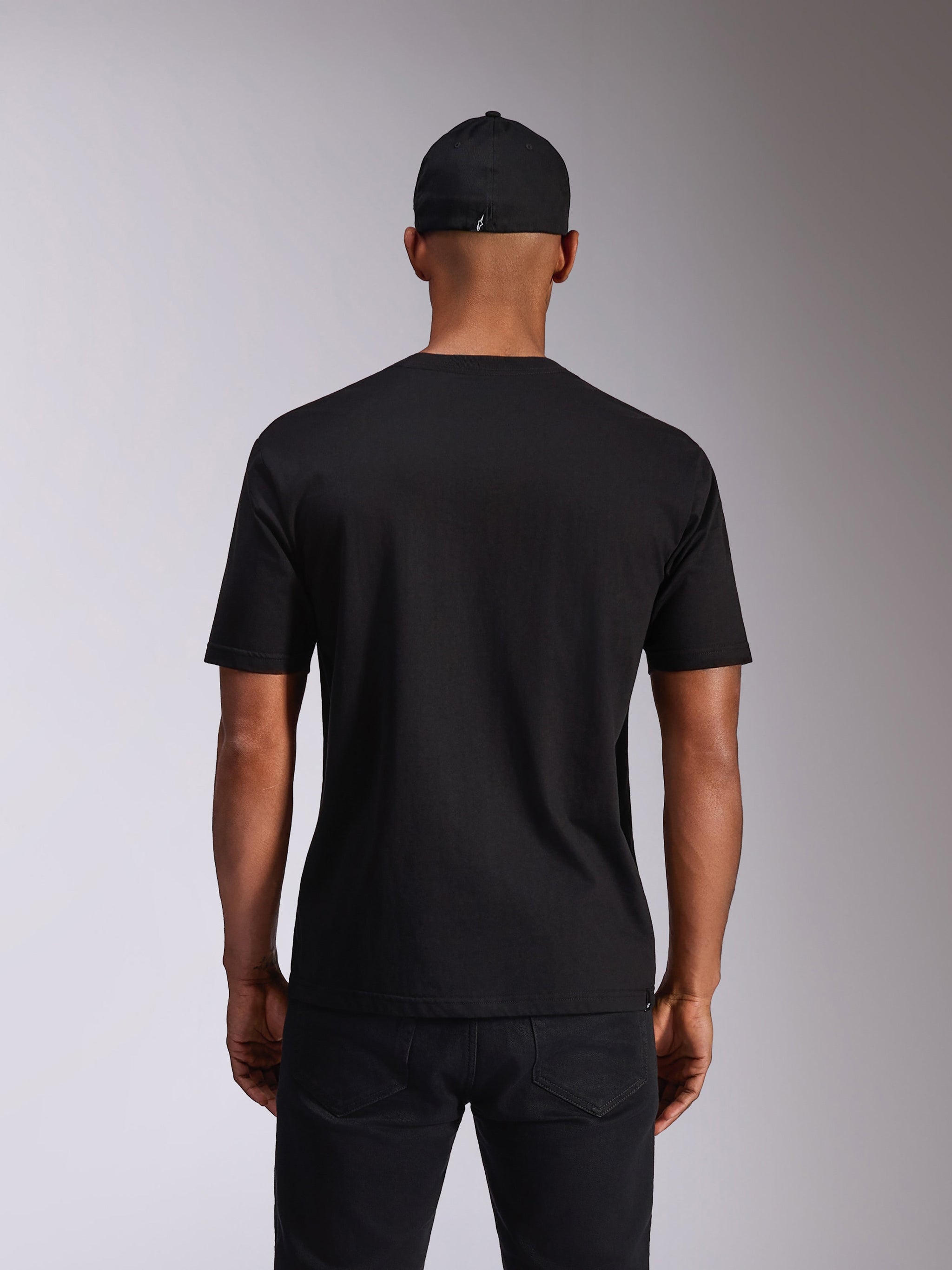 Alpinestars Ellipsoid CSF Tee, camiseta de manga corta, negra, vista trasera en modelo, combinada con una gorra negra y pantalones oscuros, ropa urbana casual inspirada en el automovilismo