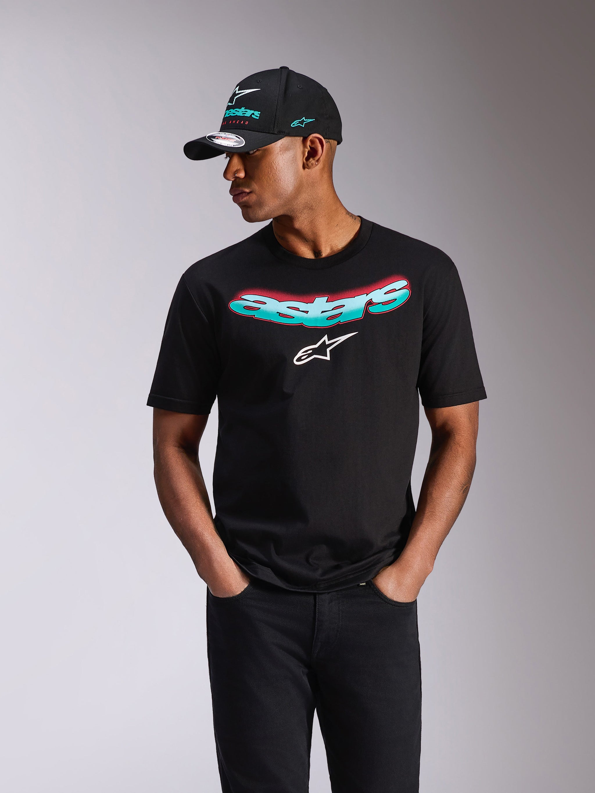Camiseta Alpinestars Ellipsoid CSF, camiseta de manga corta, negra, marca denominativa Astars en degradado verde azulado y rosa y logotipo blanco, llevada por un modelo con gorra Alpinestars a juego y vaqueros negros, estilo urbano casual de deportes de motor
