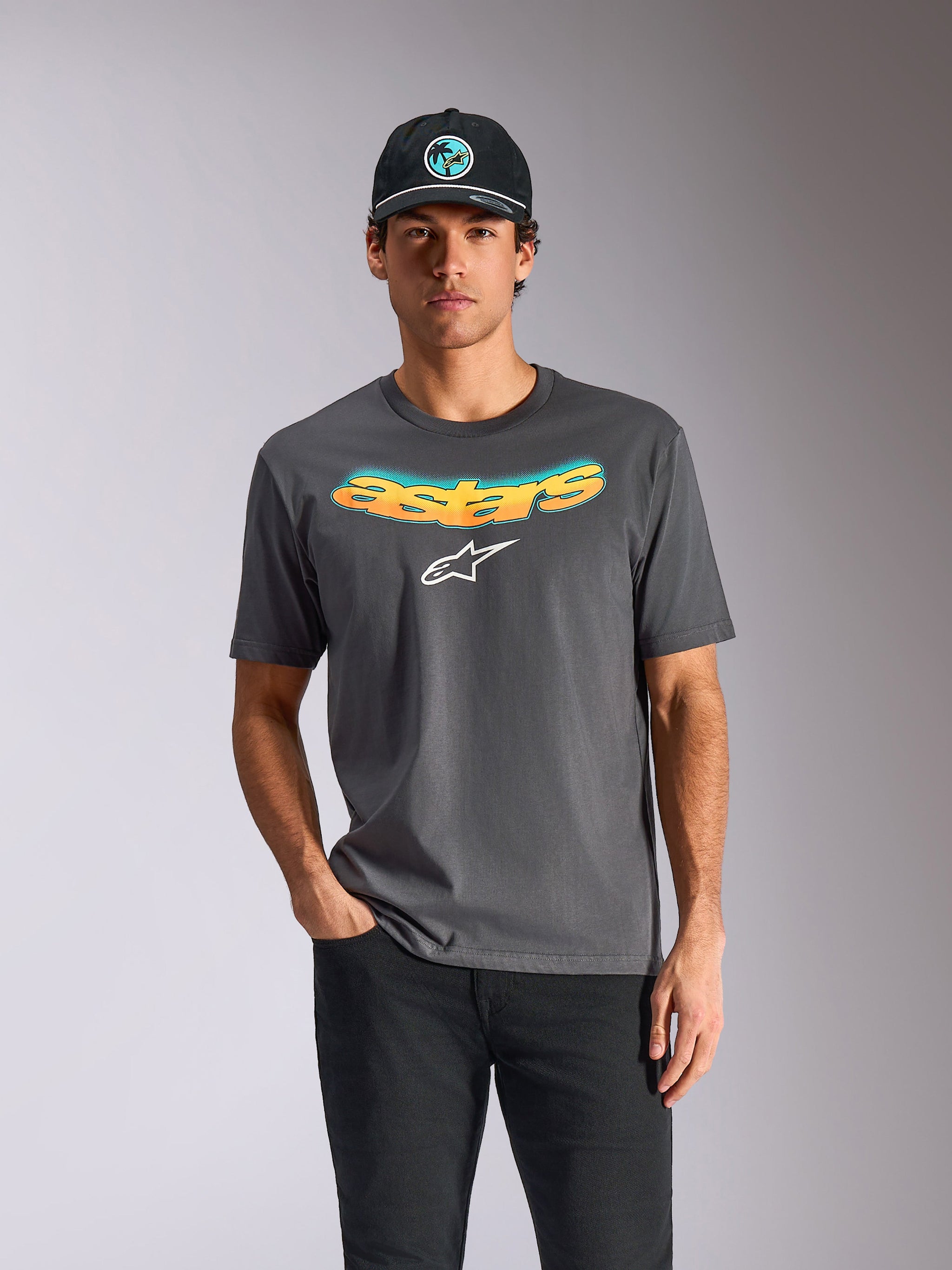 Camiseta Alpinestars Ellipsoid CSF, camiseta de manga corta en gris carbón, con la palabra "astars" curvada en degradado naranja y azul y un logotipo blanco, vestida por un modelo con gorra y pantalones negros, estilo casual de automovilismo y ropa urbana