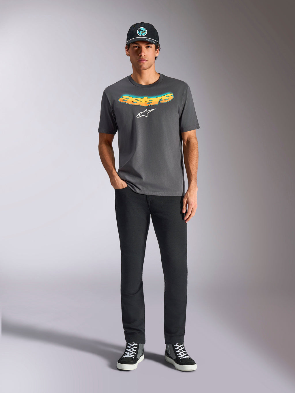 Camiseta Alpinestars Ellipsoid CSF, camiseta de manga corta, gris carbón, con palabra Astars curvada en degradado amarillo y naranja y logotipo Astars en blanco, usada por un modelo con pantalones negros y gorra de béisbol, estilo deportivo casual