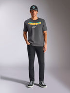 Camiseta Alpinestars Ellipsoid CSF, camiseta de manga corta, gris carbón, con palabra Astars curvada en degradado amarillo y naranja y logotipo Astars en blanco, usada por un modelo con pantalones negros y gorra de béisbol, estilo deportivo casual