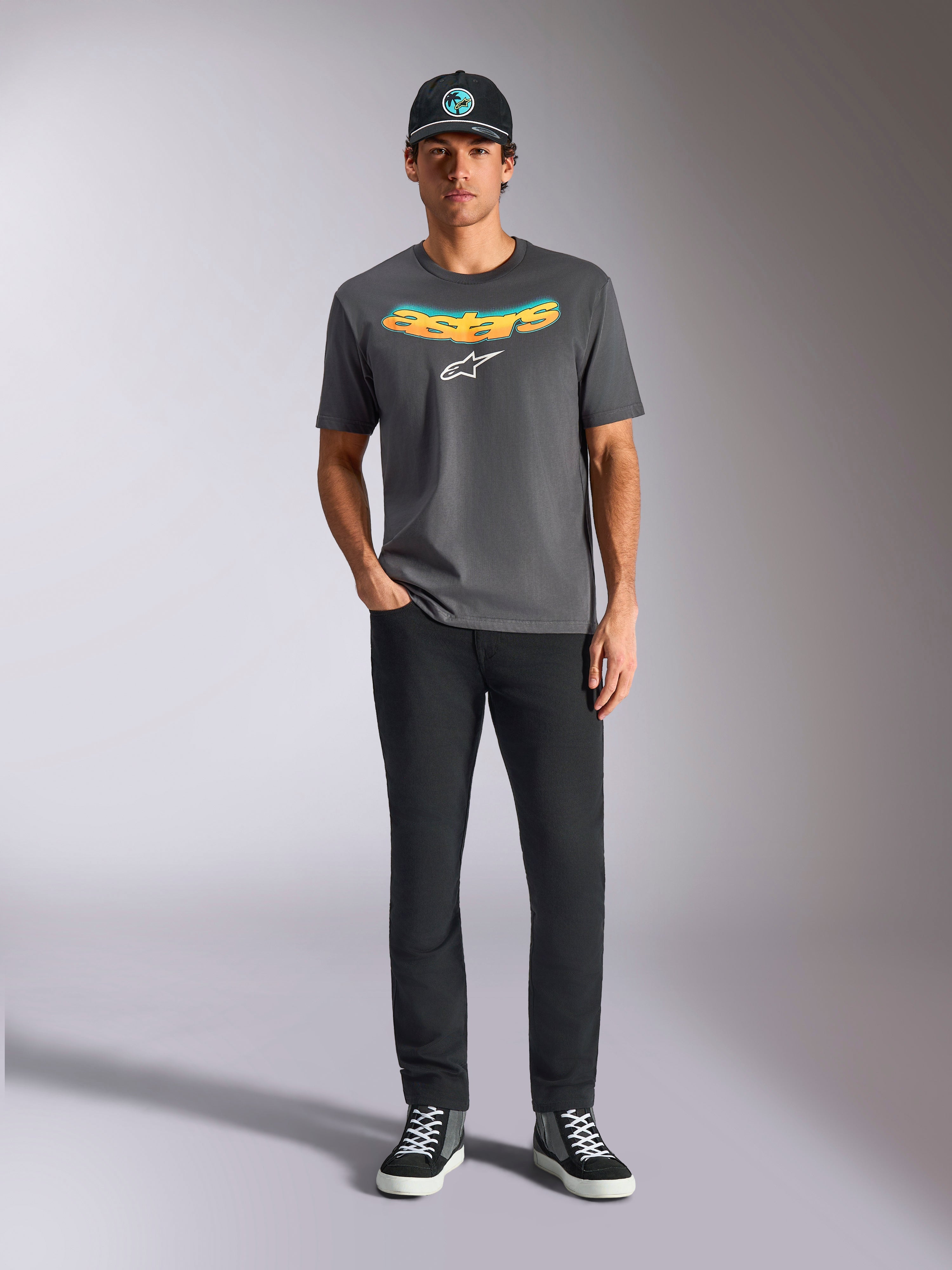 Camiseta Alpinestars Ellipsoid CSF, camiseta de manga corta, gris carbón, con palabra Astars curvada en degradado amarillo y naranja y logotipo Astars en blanco, usada por un modelo con pantalones negros y gorra de béisbol, estilo deportivo casual