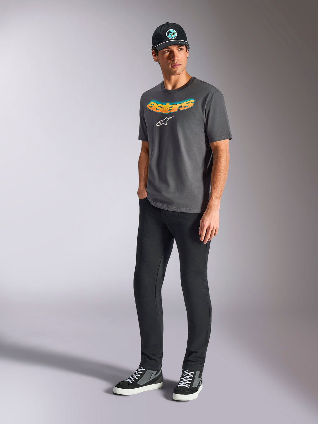 Camiseta Alpinestars Ellipsoid CSF, camiseta de manga corta, gris carbón, con la palabra astars curvada en degradado de color y logo blanco, usada por un modelo con pantalones negros y gorra, estilo casual motorsport streetwear