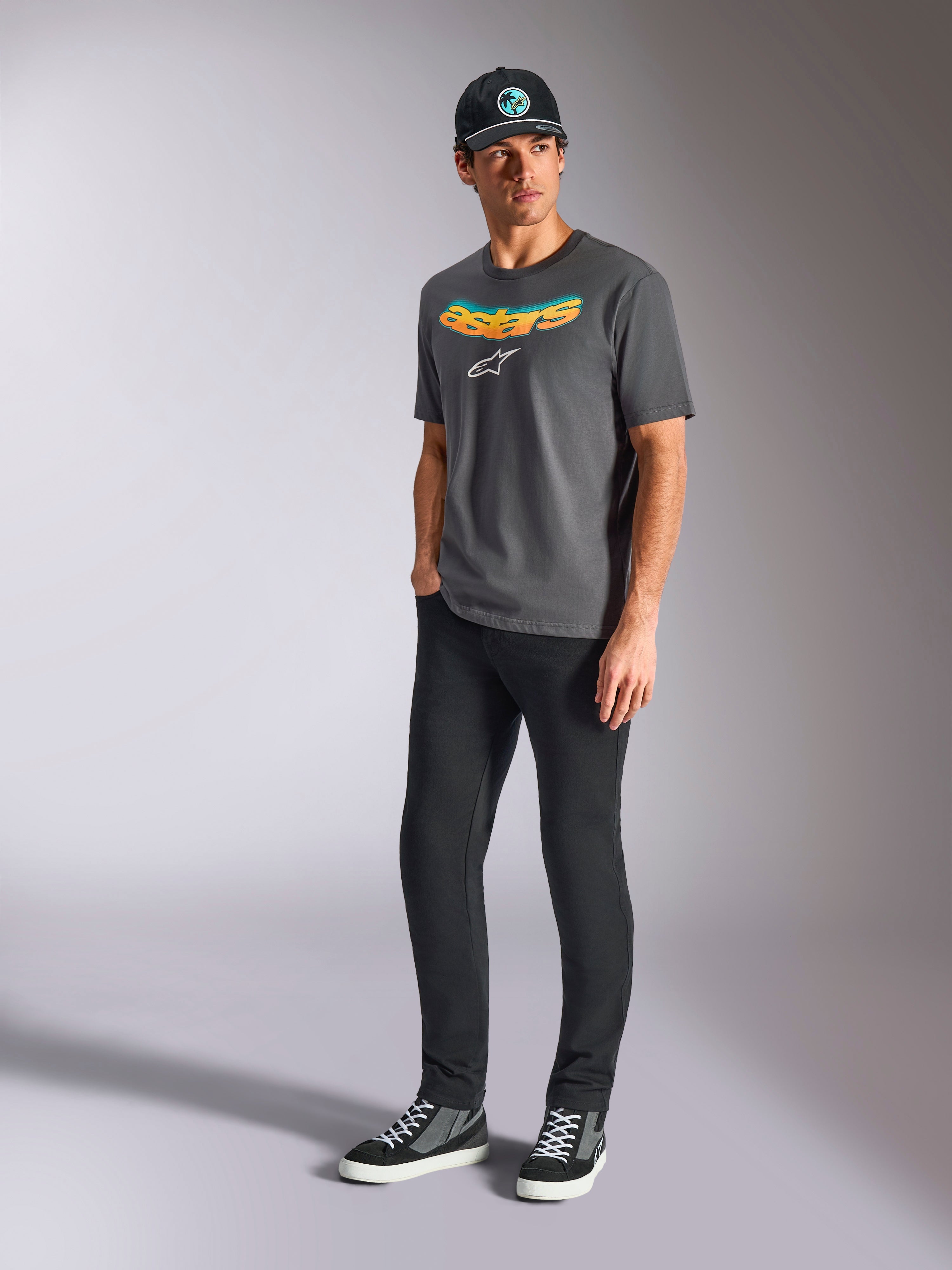 Camiseta Alpinestars Ellipsoid CSF, camiseta de manga corta, gris carbón, con la palabra astars curvada en degradado de color y logo blanco, usada por un modelo con pantalones negros y gorra, estilo casual motorsport streetwear