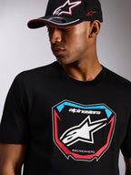 Alpinestars Plate CSF Tee, camiseta de manga corta, negra, con un gran gráfico frontal de placa de MX, logo de estrella y detalles en rojo y azul, combinada con una gorra Alpinestars negra a juego, estilo casual motorsport streetwear