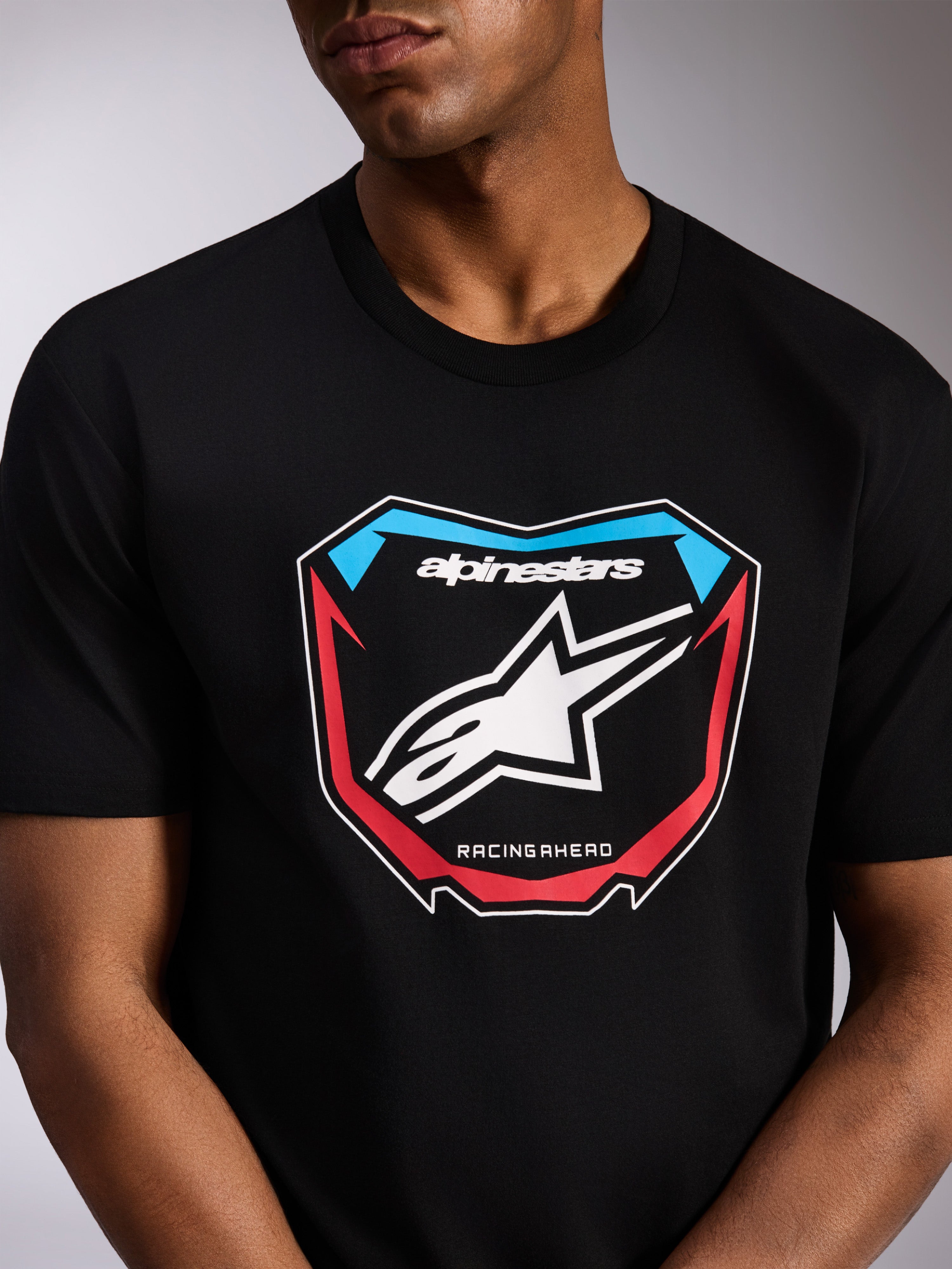 Alpinestars Plate CSF Tee, camiseta de manga corta, negra, gráfico frontal de placa de motocross con logotipo Astars blanco, detalles en rojo y azul, y texto 'RACING AHEAD', ropa urbana casual inspirada en el motociclismo