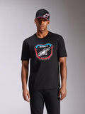 Alpinestars Plate CSF Tee, camiseta de manga corta, negra, con gráfico frontal de placa de MX con logotipo blanco y detalles en rojo y azul, usada por un modelo con gorra negra a juego, ropa urbana casual de automovilismo
