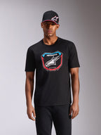 Alpinestars Plate CSF Tee, camiseta de manga corta, negra, con gráfico frontal de placa de MX con logotipo blanco y detalles en rojo y azul, usada por un modelo con gorra negra a juego, ropa urbana casual de automovilismo