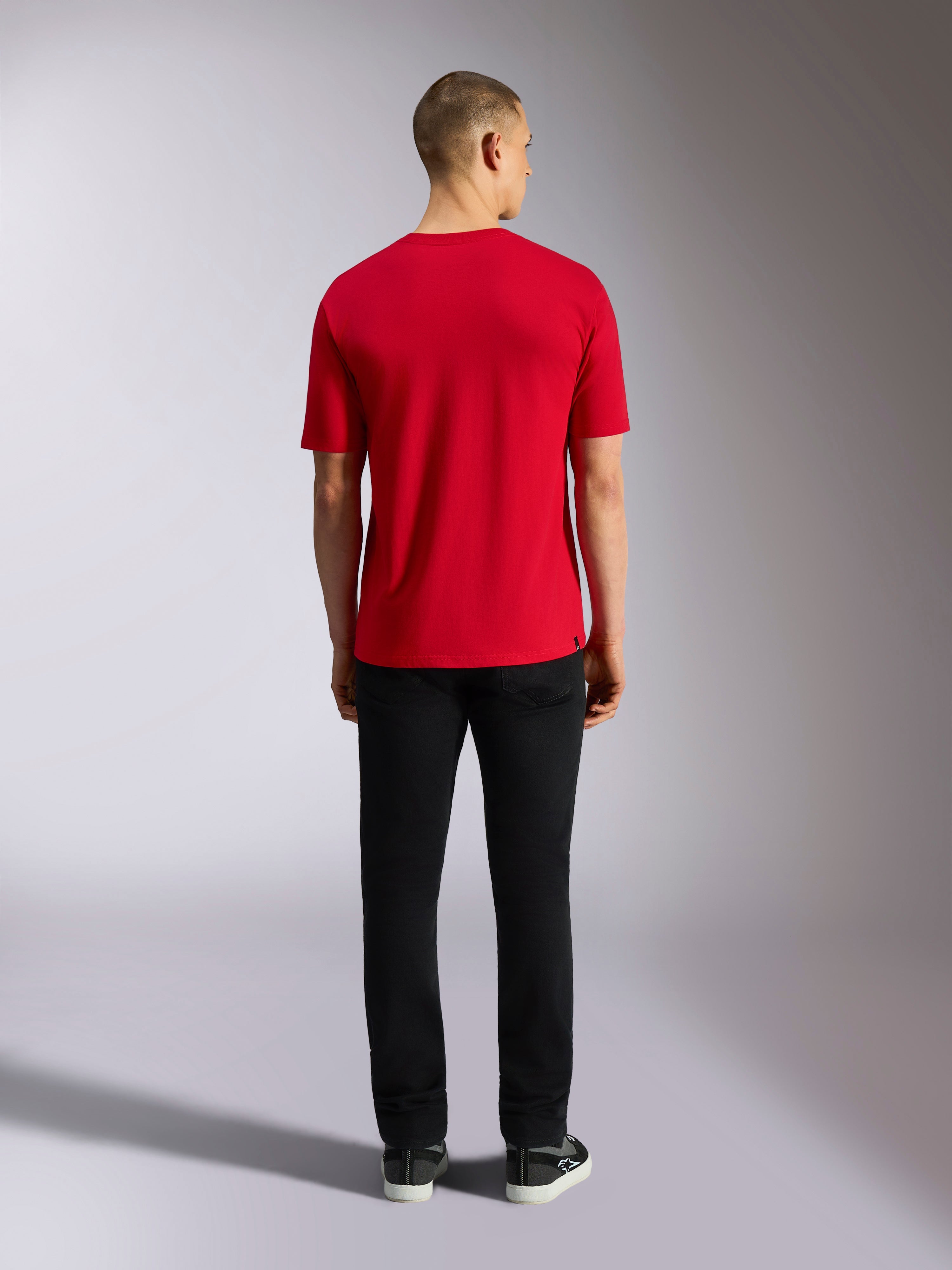 Alpinestars Plate CSF Tee, camiseta de manga corta, roja, vista posterior del modelo con pantalones negros, ropa deportiva casual inspirada en el automovilismo