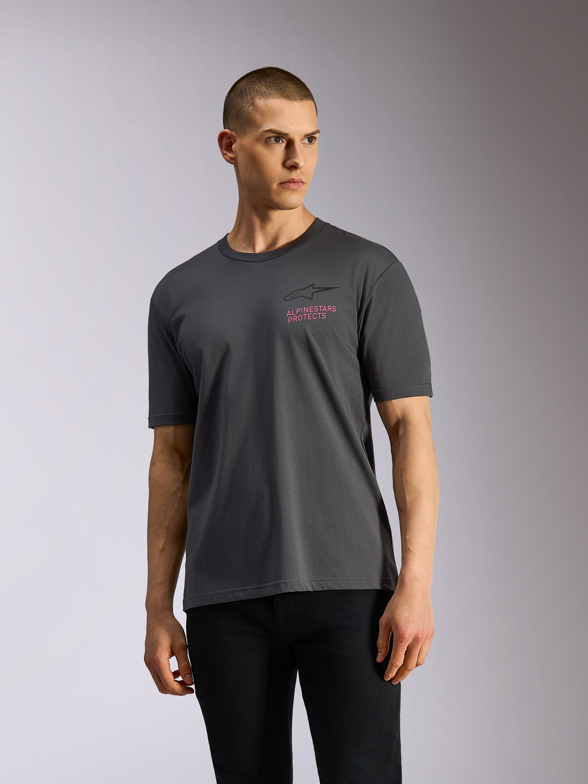 Alpinestars Pop Helmet CSF Tee, camiseta de manga corta, gris carbón, lucida por un modelo, con el logo Astars en negro y el texto 'ALPINESTARS PROTECTS' en rosa en el pecho, estilo casual motorsport