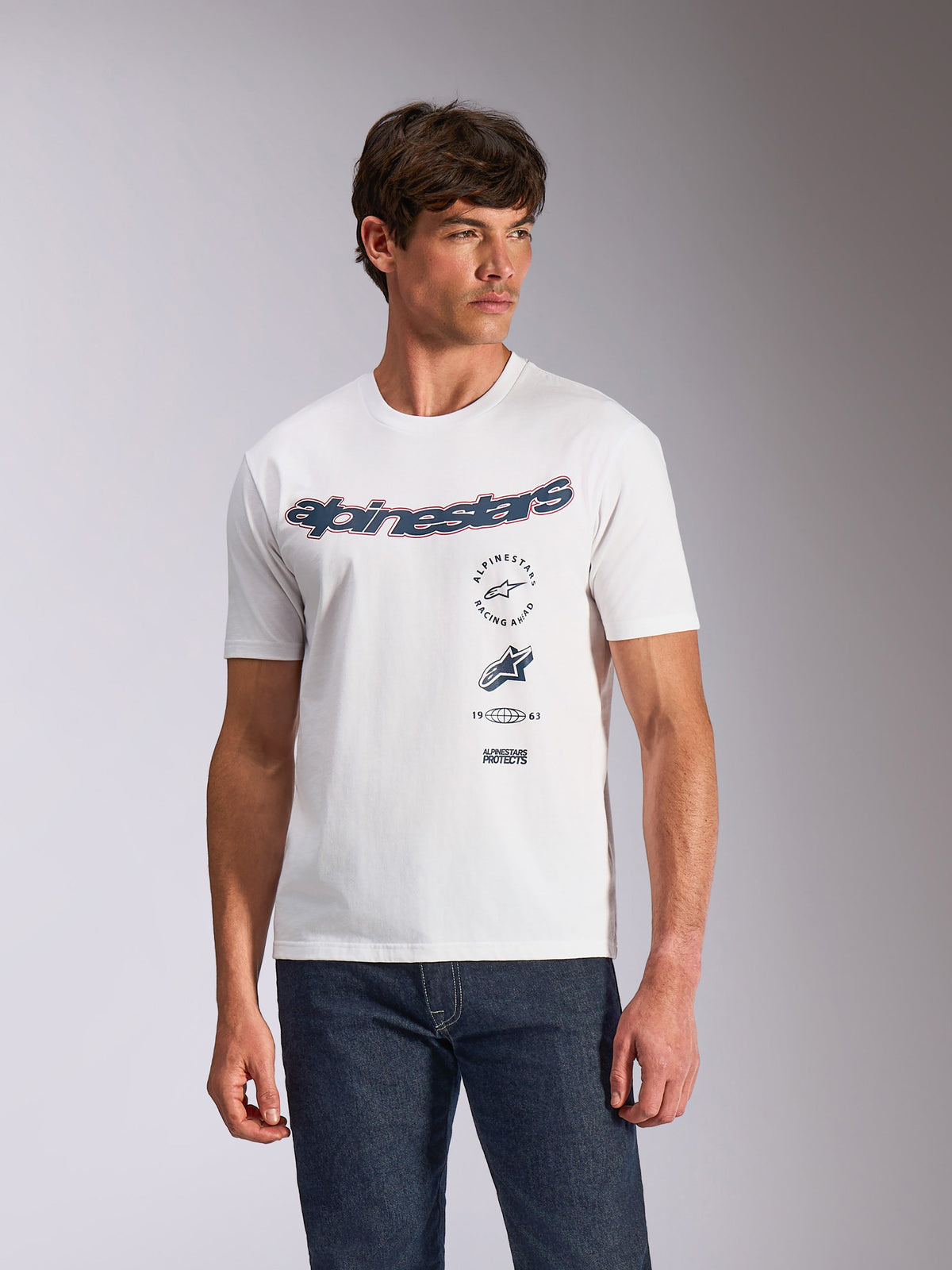 Camiseta Alpinestars Abus CSF, camiseta de manga corta, blanca, logotipo curvado de Alpinestars con múltiples gráficos apilados en el pecho, lucida por un modelo con vaqueros oscuros, estilo urbano casual de inspiración motorsport