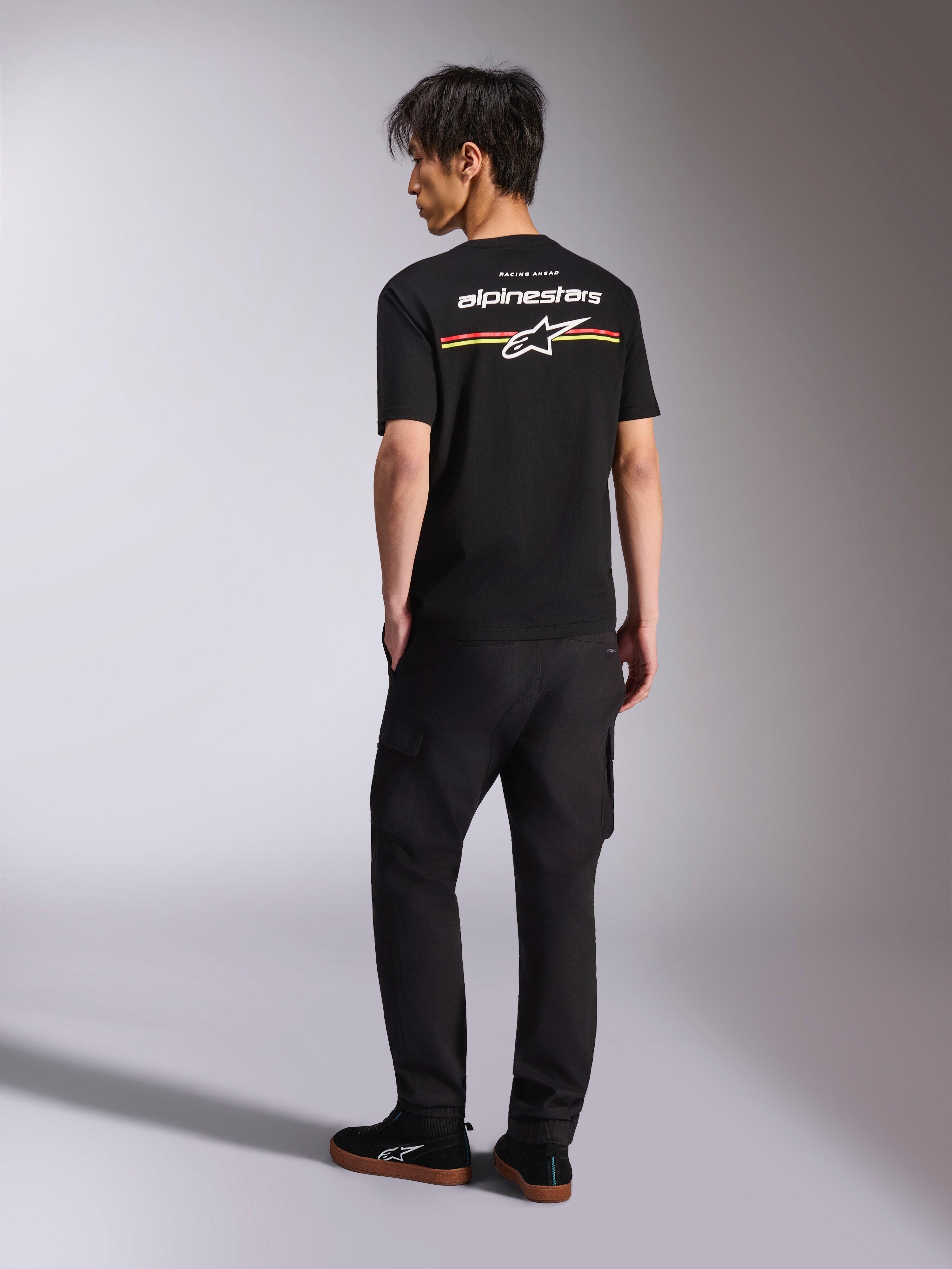 Alpinestars Lanes CSF Tee, camiseta de manga corta negra, vista posterior con el logotipo de Alpinestars en blanco y gráficos de rayas rojas, naranjas y amarillas, ropa urbana casual de automovilismo combinada con pantalones cargo negros