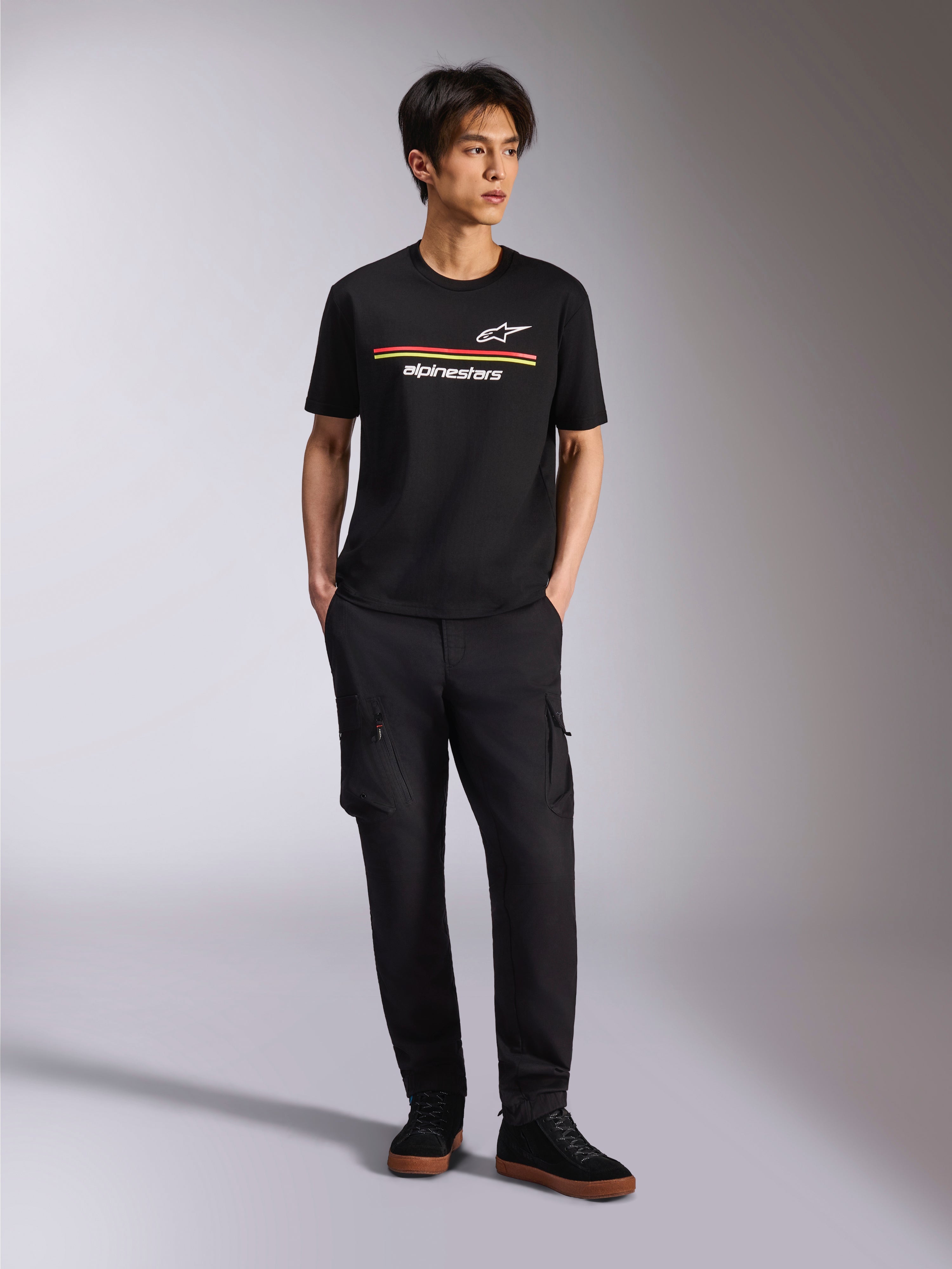 Modelo vistiendo la camiseta Alpinestars Lanes CSF de manga corta, negra, con franjas rojas y amarillas y logotipos de Astars y Alpinestars, combinada con pantalones cargo negros y zapatillas, ropa urbana casual de estilo motorsport