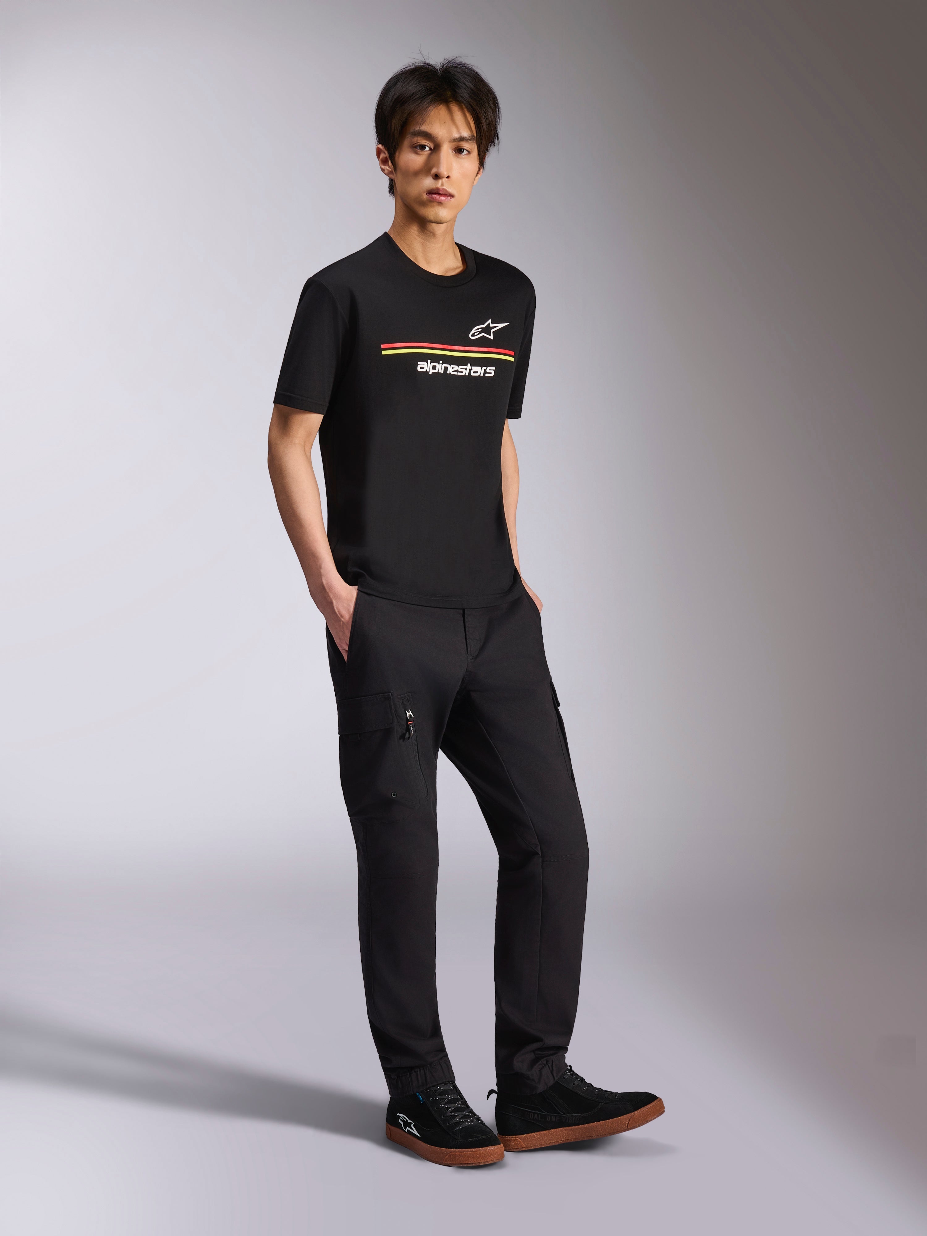 Alpinestars Lanes CSF Tee, camiseta de manga corta, negra, logotipos de rayas multicolores de Astars y Alpinestars en el pecho, usada por un modelo con pantalones cargo negros, estilo casual de ropa de calle inspirado en el automovilismo