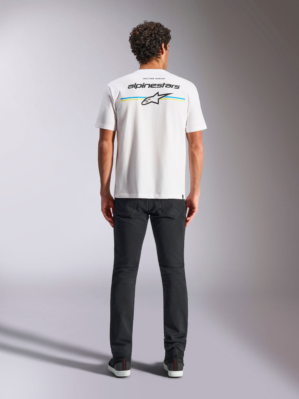 Camiseta Alpinestars Lanes CSF, camiseta de manga corta, blanca, vista posterior en modelo con el logotipo de Alpinestars y la estrella Astars sobre franjas horizontales azules y amarillas, ropa urbana casual de estilo motorsport