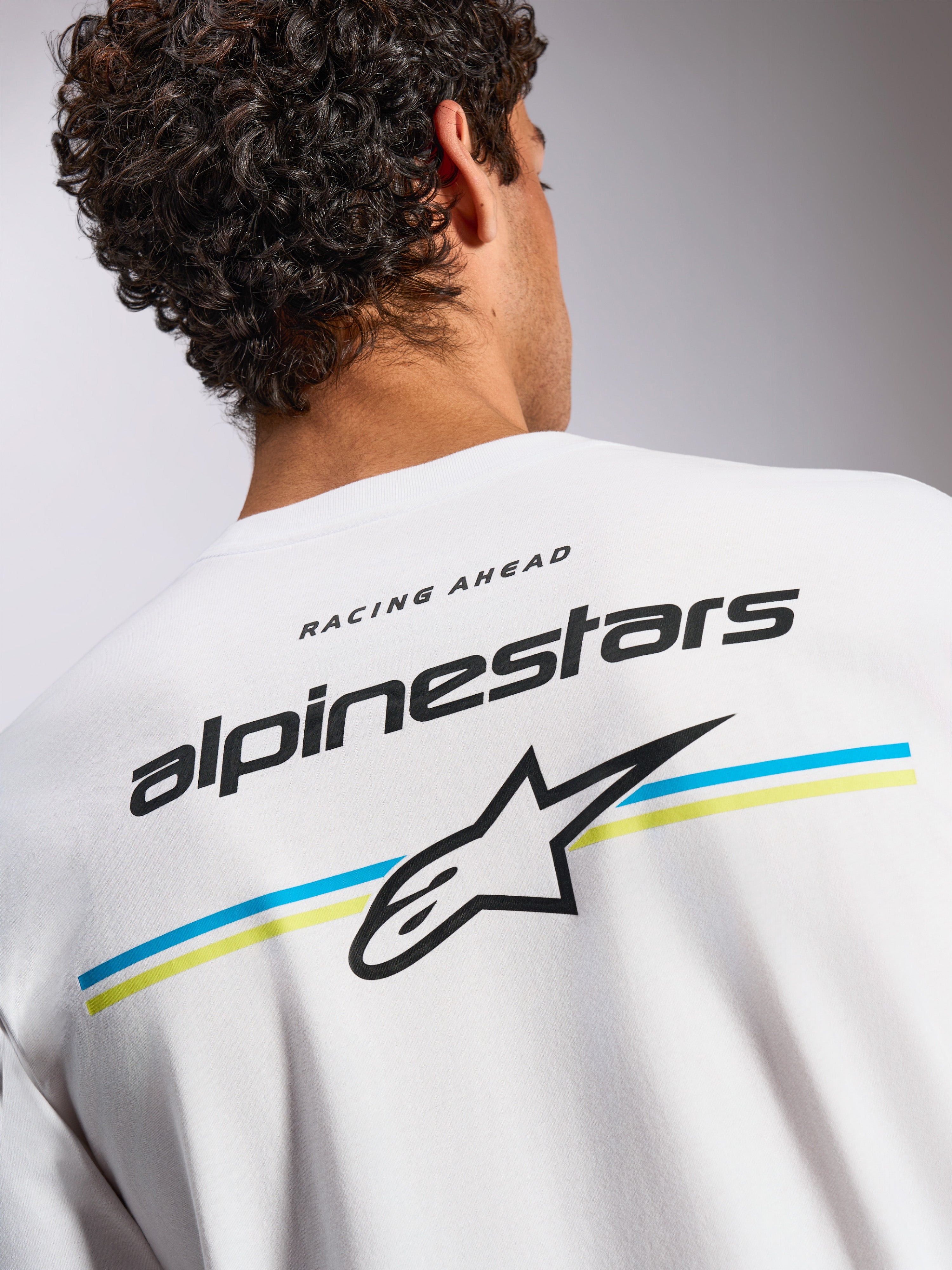 Alpinestars Lanes CSF Tee, camiseta de manga corta, blanca, gráfico en la espalda con el nombre Alpinestars y el logotipo Astars sobre rayas azules y amarillas, ropa urbana casual inspirada en el automovilismo
