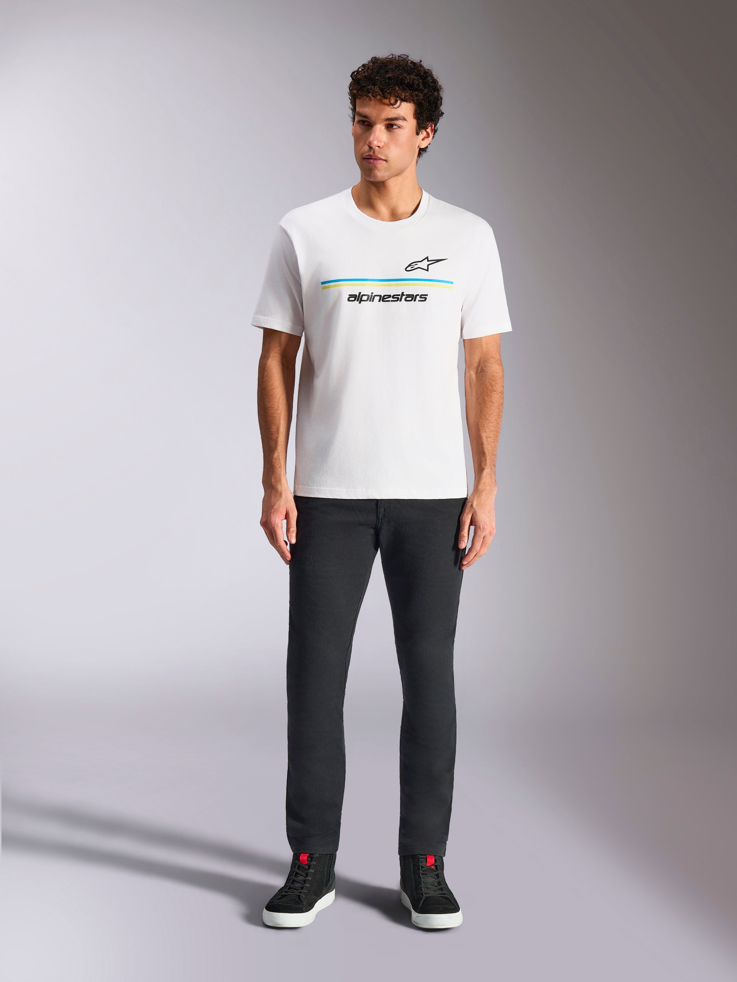 Alpinestars Lanes CSF Tee, camiseta de manga corta, blanca, gráfico de logotipo con franjas azules y amarillas, usada por un modelo con pantalones negros y zapatillas, ropa urbana casual de automovilismo