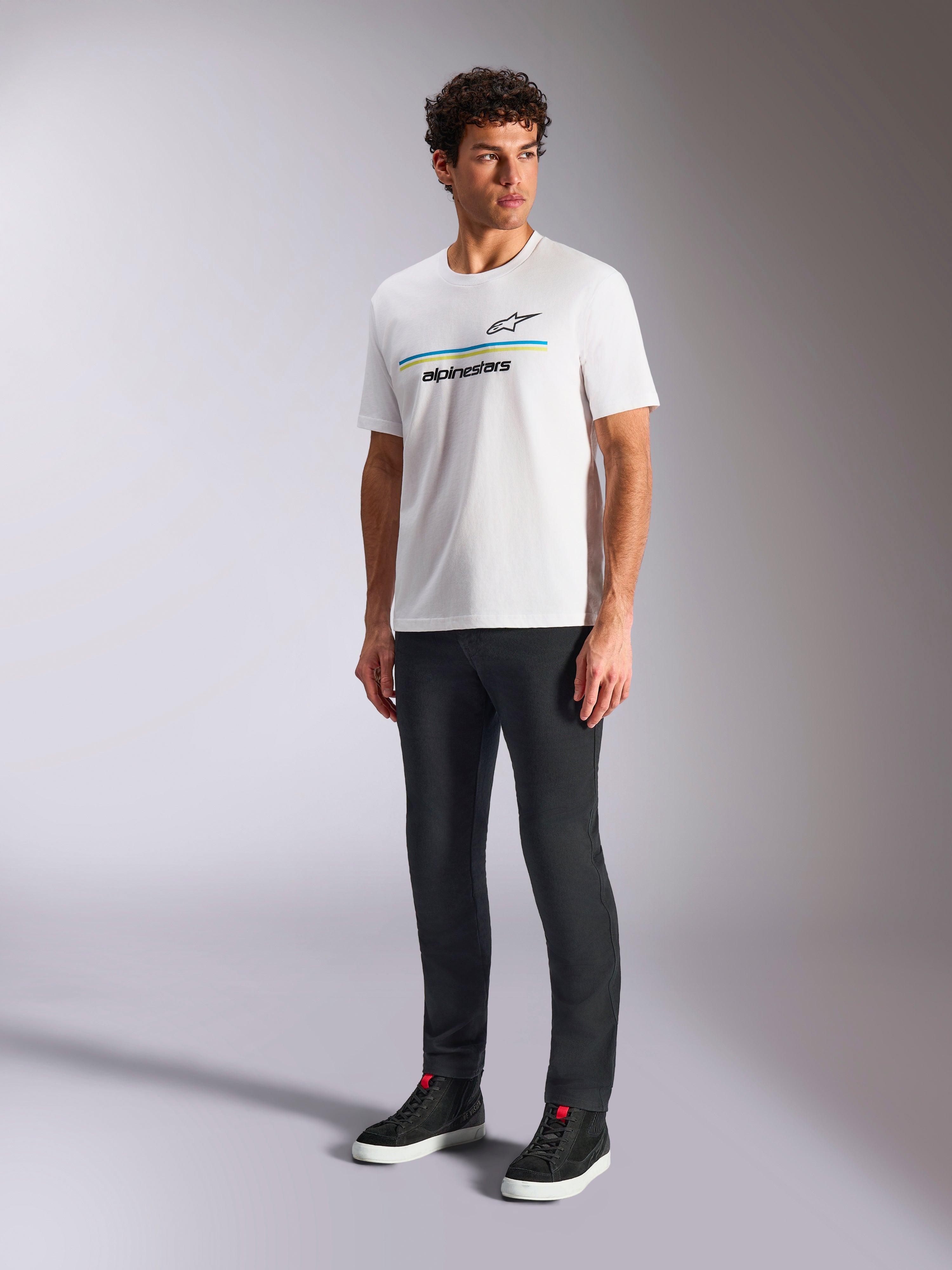 Camiseta Alpinestars Lanes CSF, camiseta blanca de manga corta, vestida por un modelo con pantalones negros ajustados y zapatillas altas negras, con rayas horizontales azules y amarillas y el logotipo de Alpinestars en el pecho, estilo urbano casual inspirado en el automovilismo
