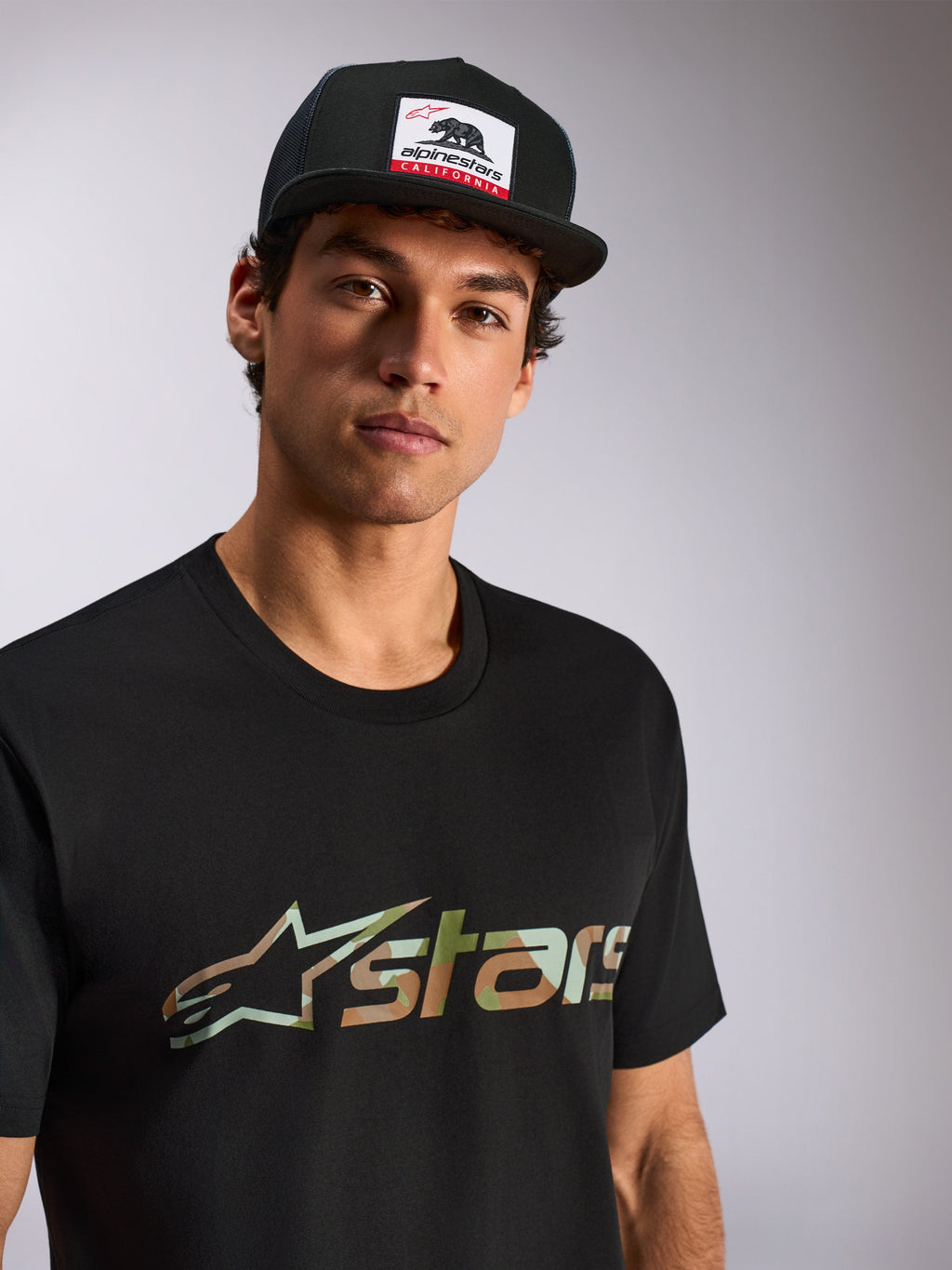 Camiseta Alpinestars Verdure Camo CSF, de manga corta, negra, logo Astars con relleno de camuflaje, usada por modelo masculino con gorra trucker Alpinestars California negra, estilo casual motorsport streetwear