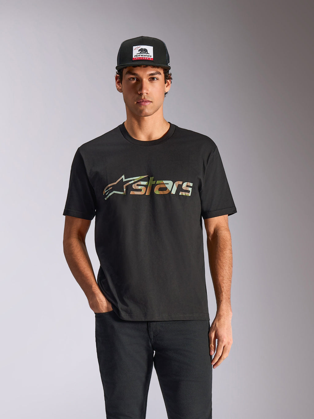 Camiseta Alpinestars Verdure Camo CSF, camiseta de manga corta negra, usada por un modelo con gorra y pantalones negros, con el logo Astars frontal con relleno de camuflaje, estilo streetwear casual de automovilismo