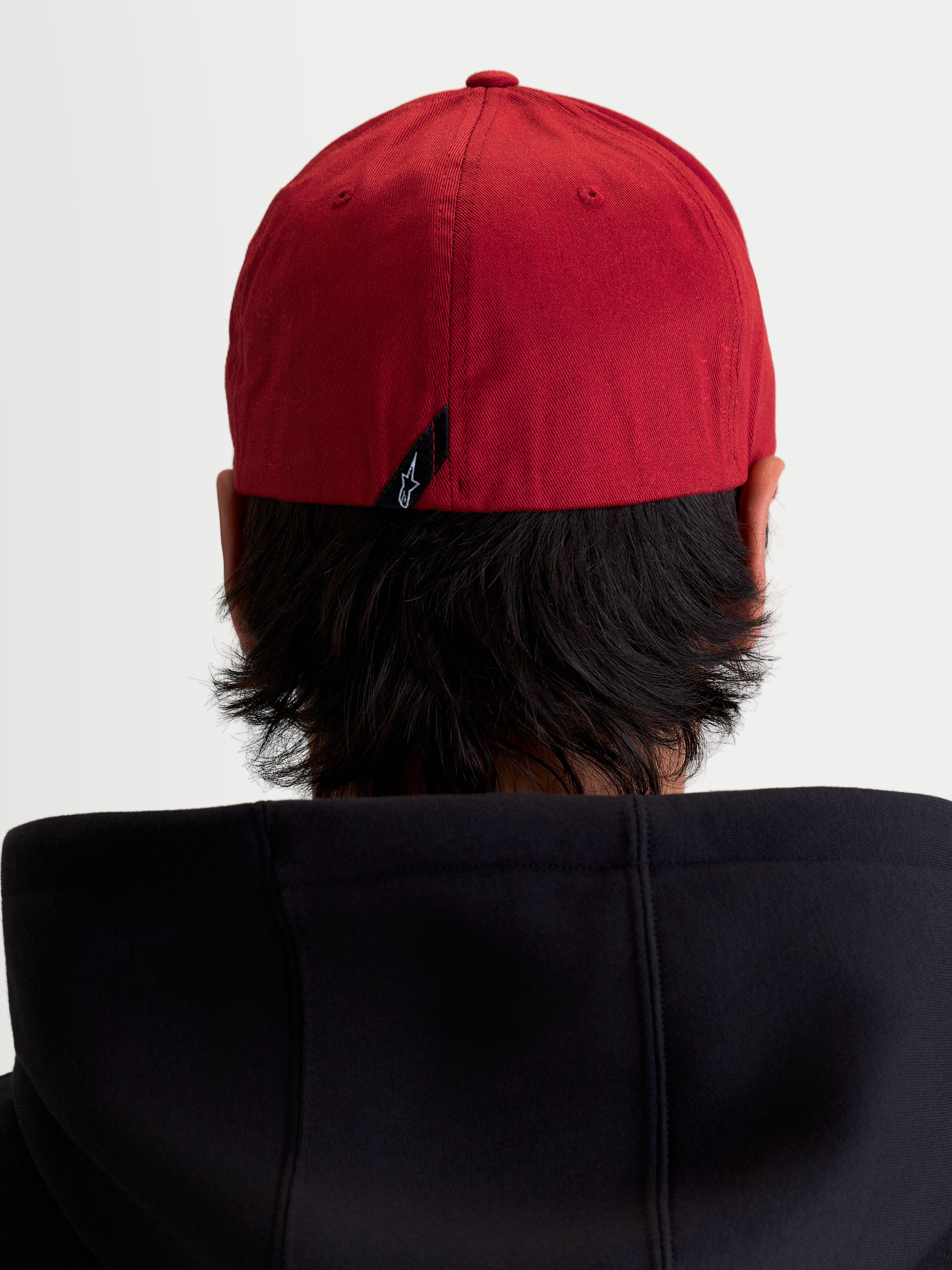 Alpinestars Gorra Pivot, gorra deportiva casual de color rojo oscuro, vista trasera sobre modelo que muestra la construcción elástica flex back y la etiqueta con el logotipo Astars