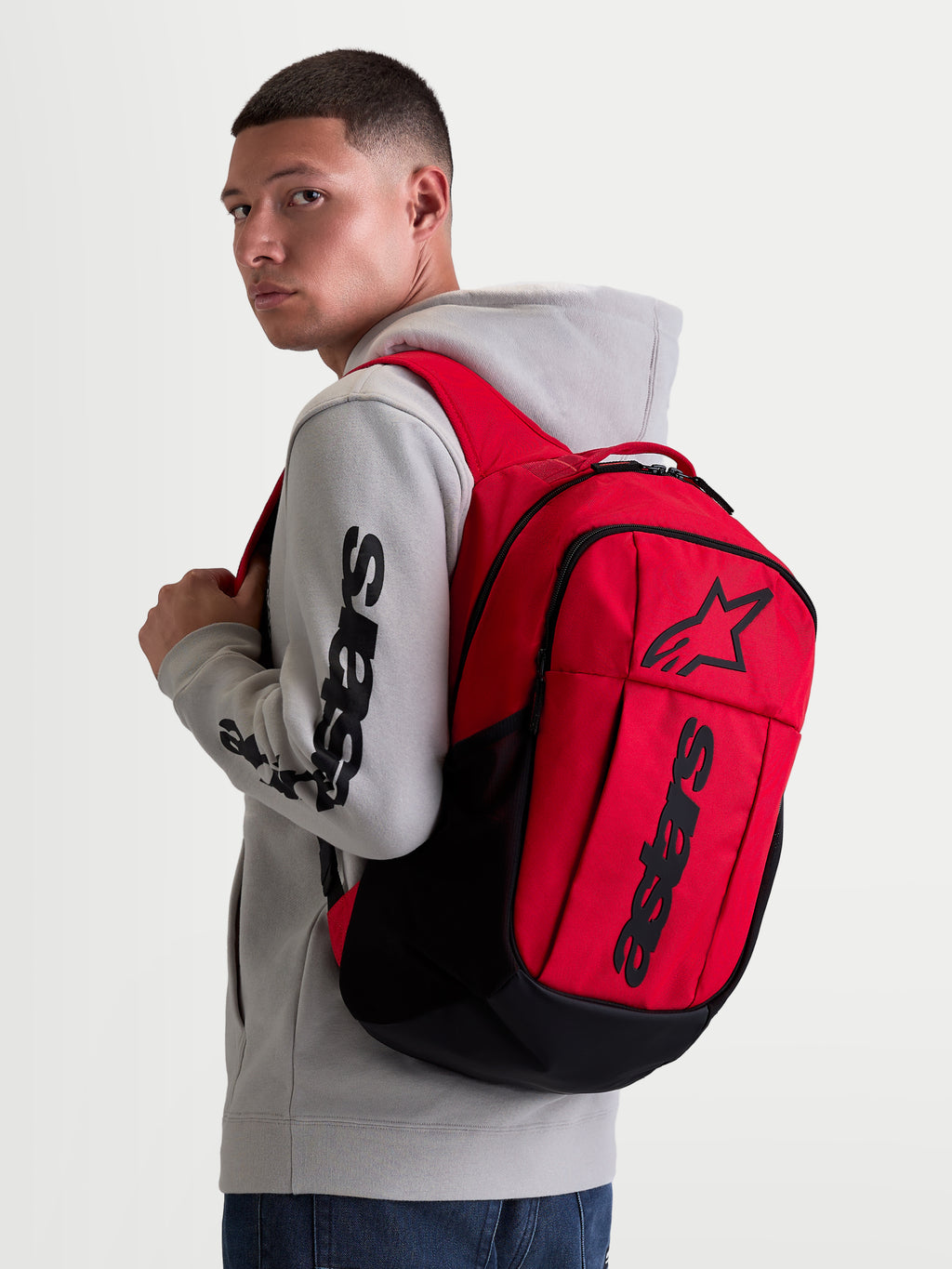 Alpinestars GFX Boss, mochila de ropa deportiva urbana de 20L, roja y negra, con construcción de poliéster 600D y logotipo Astars de silicona en relieve, mostrada en un modelo con sudadera gris.