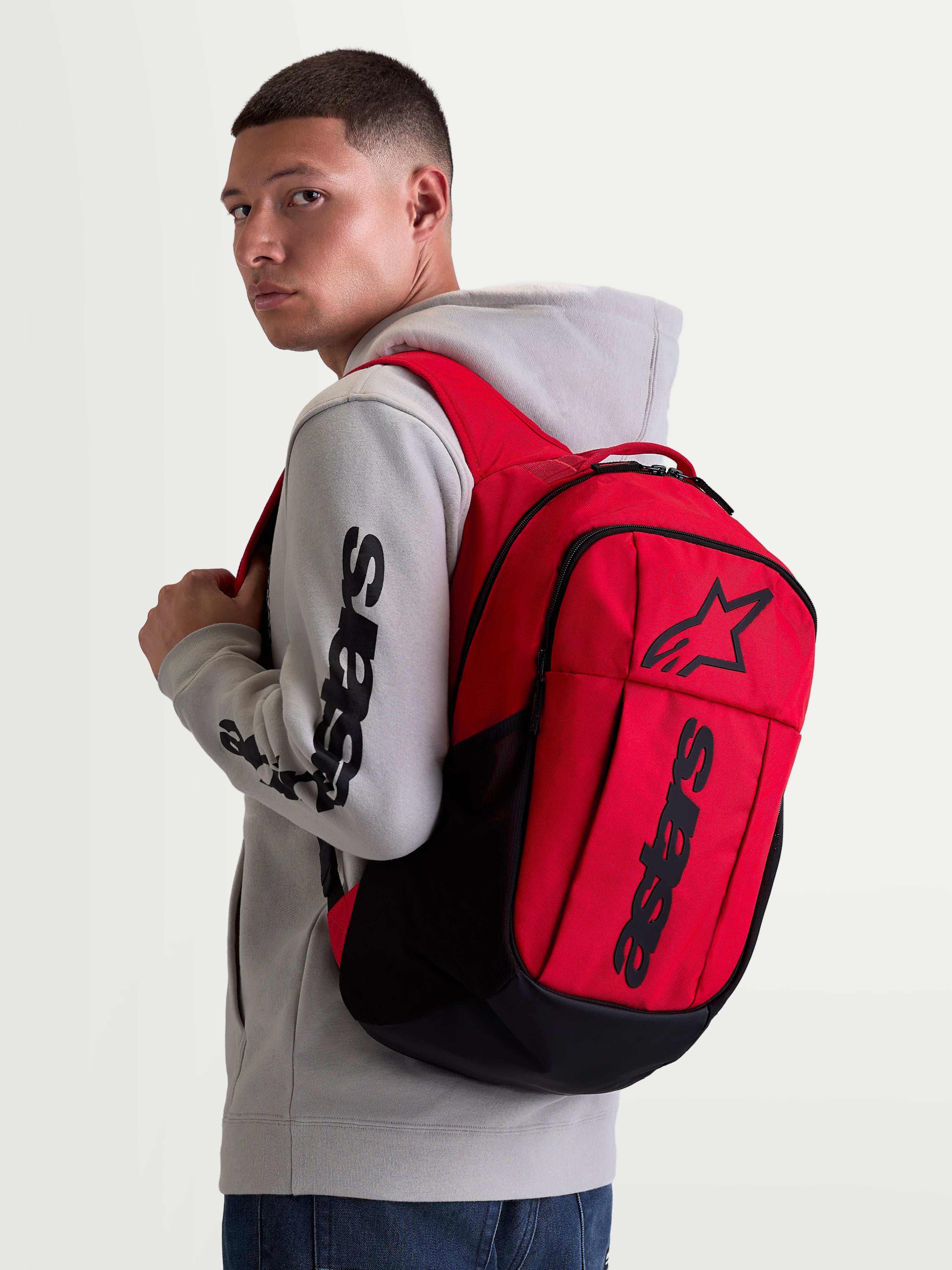 Alpinestars GFX Boss, mochila de ropa deportiva urbana de 20L, roja y negra, con construcción de poliéster 600D y logotipo Astars de silicona en relieve, mostrada en un modelo con sudadera gris.