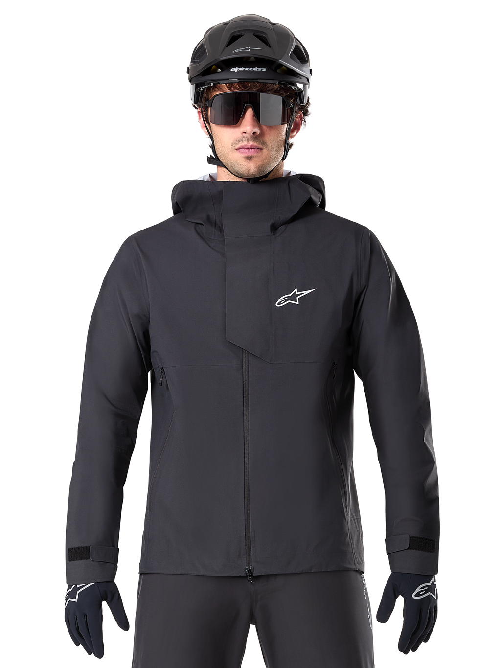 Chaqueta impermeable Alpinestars A-Dura Elite, negra, chaqueta de ciclismo de montaña, diseño de cuello alto con capucha, cremallera frontal con solapa protectora en el pecho y logotipo blanco, usada con casco negro y guantes de ciclismo, protección técnica contra la intemperie para rodar por senderos